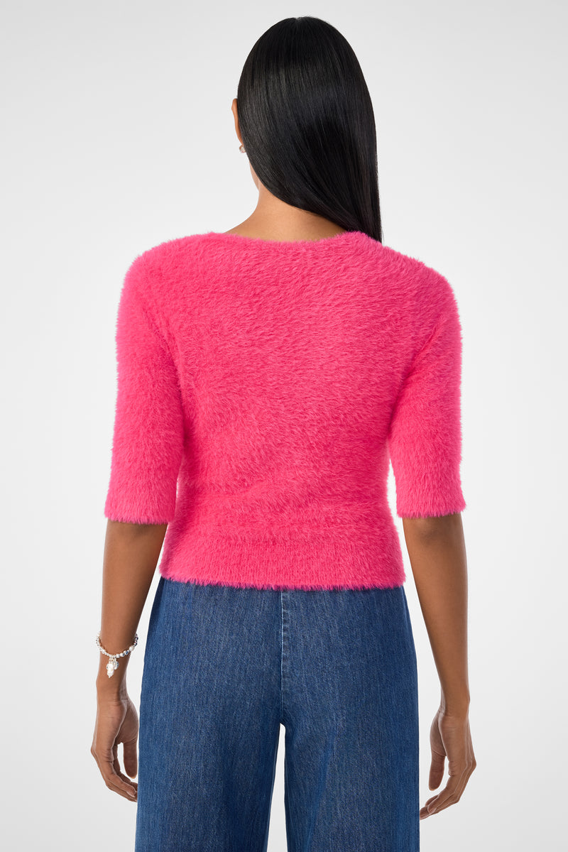 A woman with straight, dark hair wears the Maliah Fuzzy Sweater in bright pink and blue jeans. She faces away from the camera, showing a bracelet on her left wrist against a plain white background.