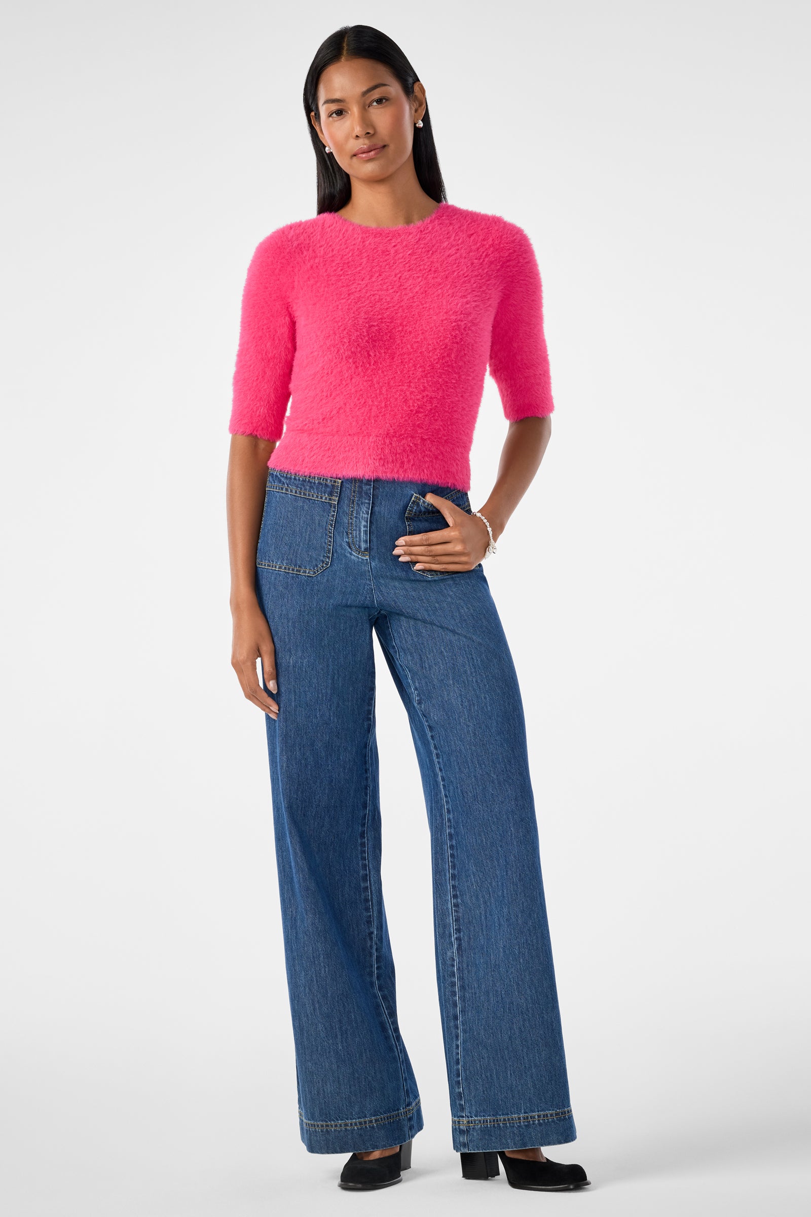 A woman with long dark hair stands against a plain background wearing the bright pink Maliah Fuzzy Sweater, high-waisted wide-leg blue jeans with large pockets, and black shoes, her expression relaxed.