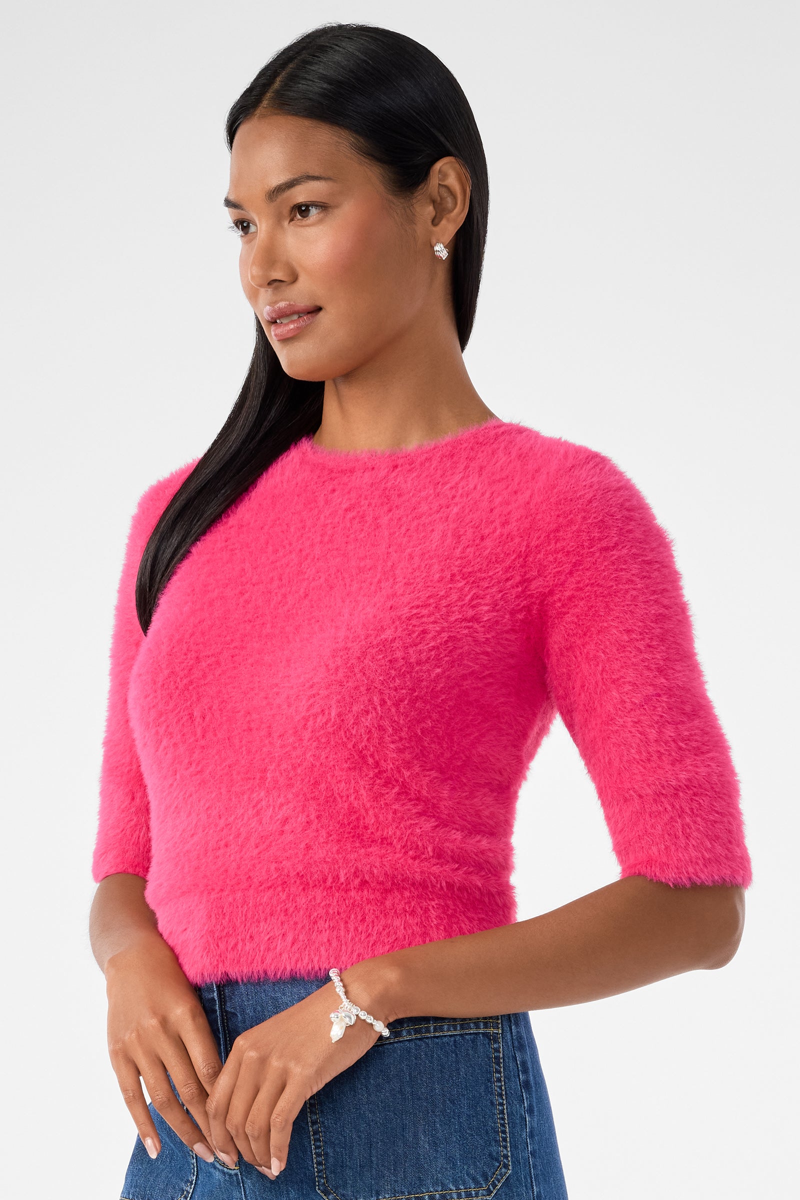 A woman with long black hair wears the Maliah Fuzzy Sweater in bright pink, paired with blue jeans. She looks to the side, smiling slightly, styled with stud earrings and a bracelet against a plain white background.