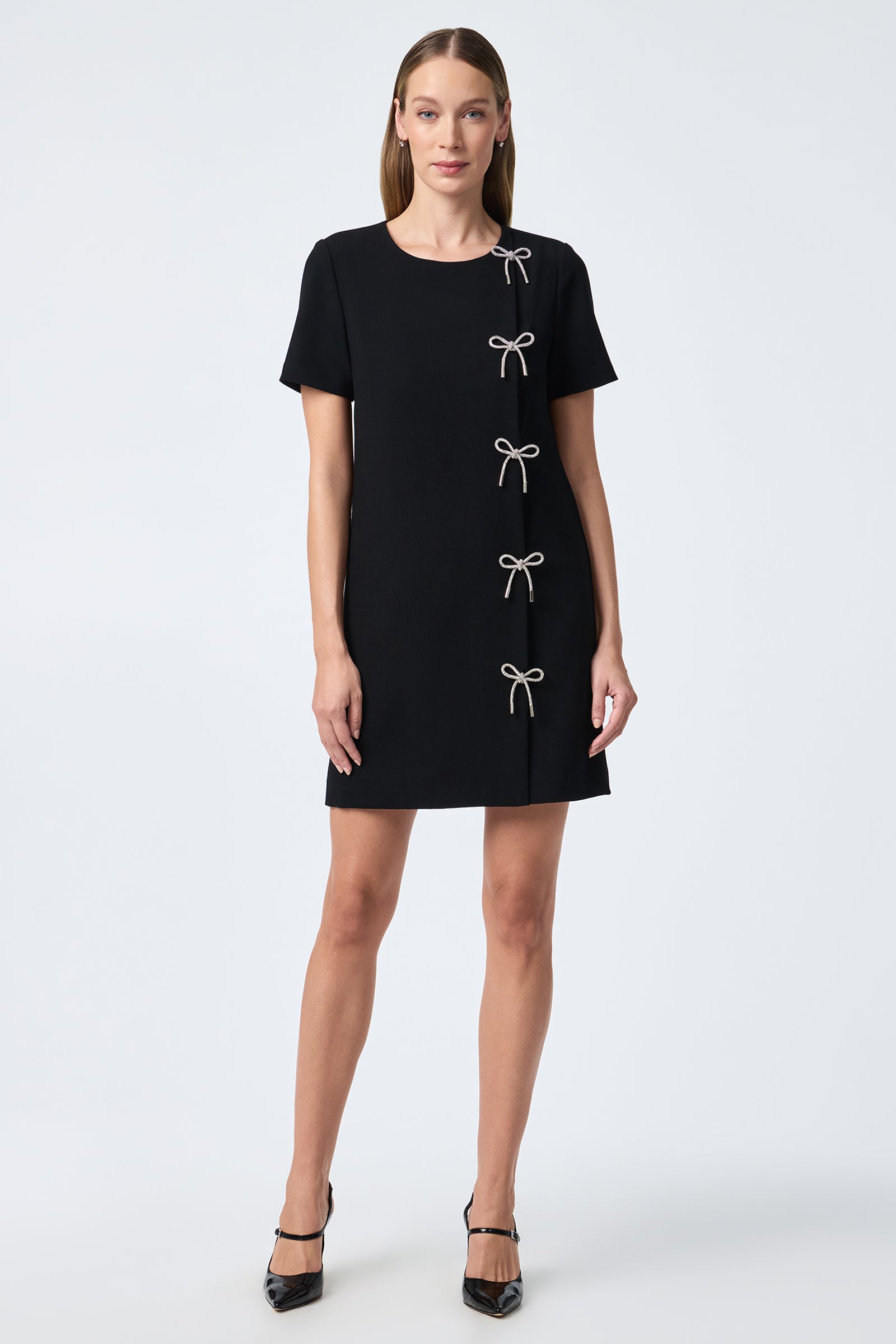 A woman wears the Diamonds Crystal Bows Sheath Dress—a short-sleeved, knee-length black dress featuring four crystal bow accents—paired with black heels against a plain light background.