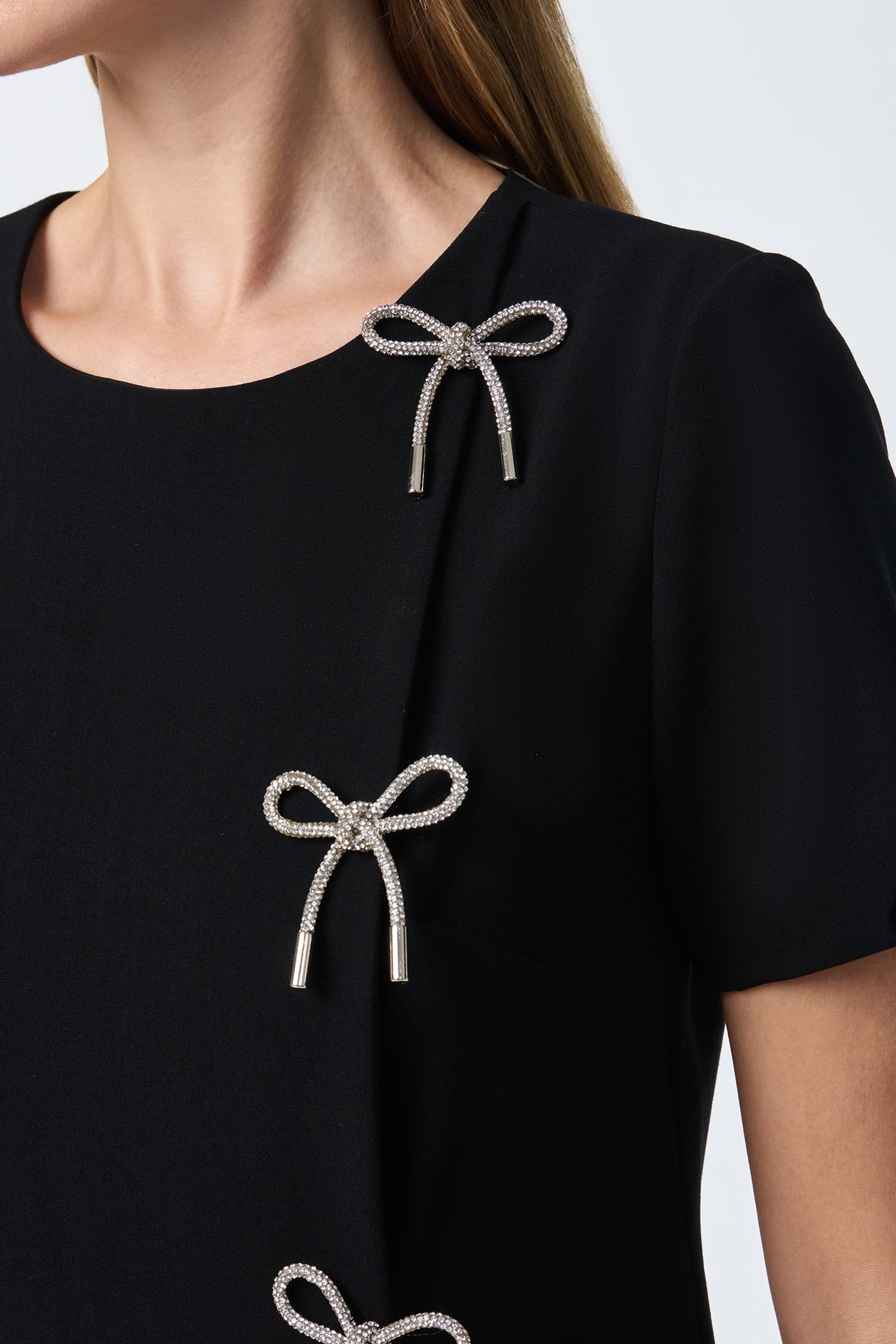 A woman wears the Diamonds Crystal Bows Sheath Dress, featuring a black fabric with silver bow-shaped embellishments on the front, short sleeves, and a round neckline.