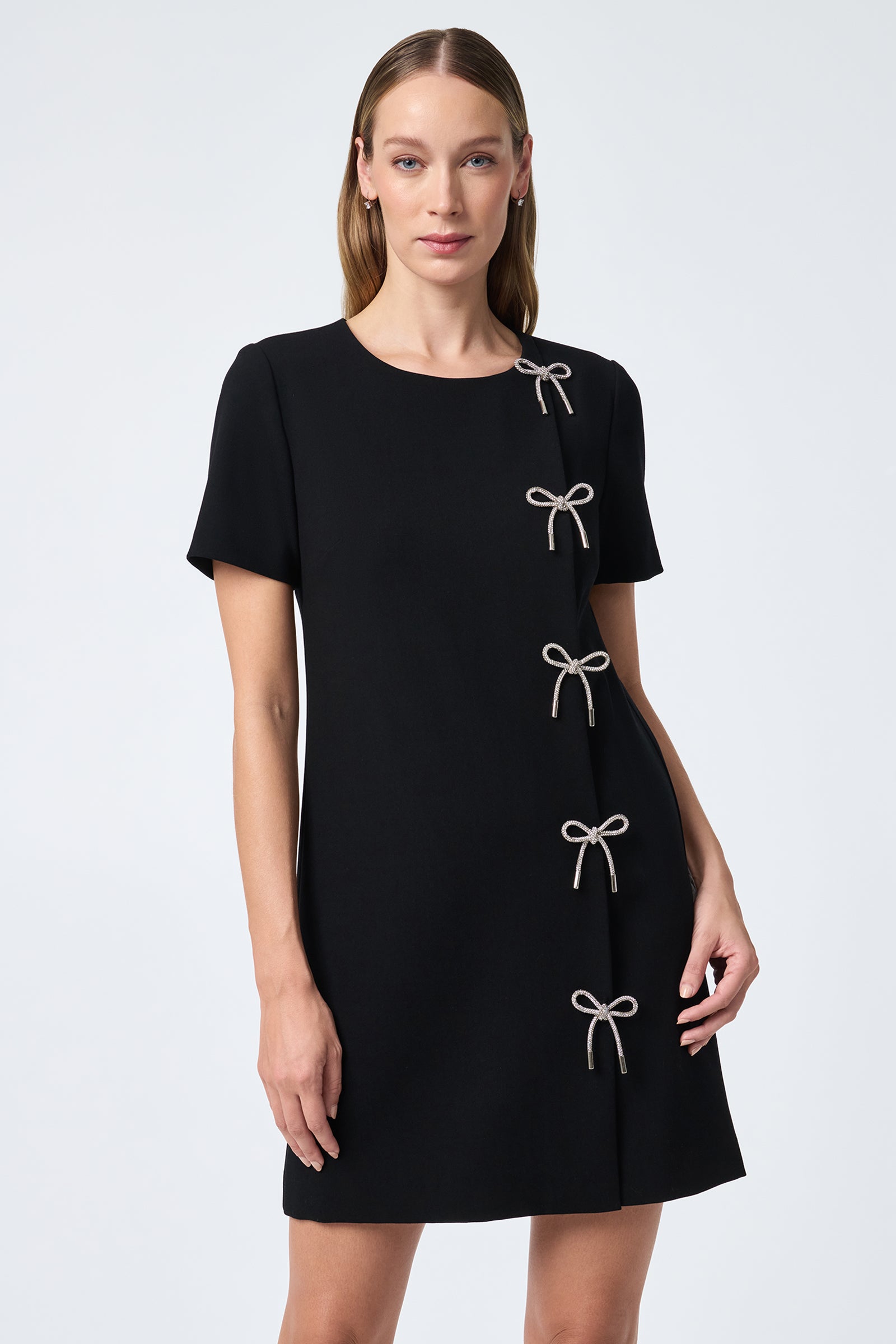 A woman with straight, light brown hair models the Diamonds Crystal Bows Sheath Dress—a short-sleeved black dress with a straight silhouette and five diamond crystal bows arranged vertically along the right front.