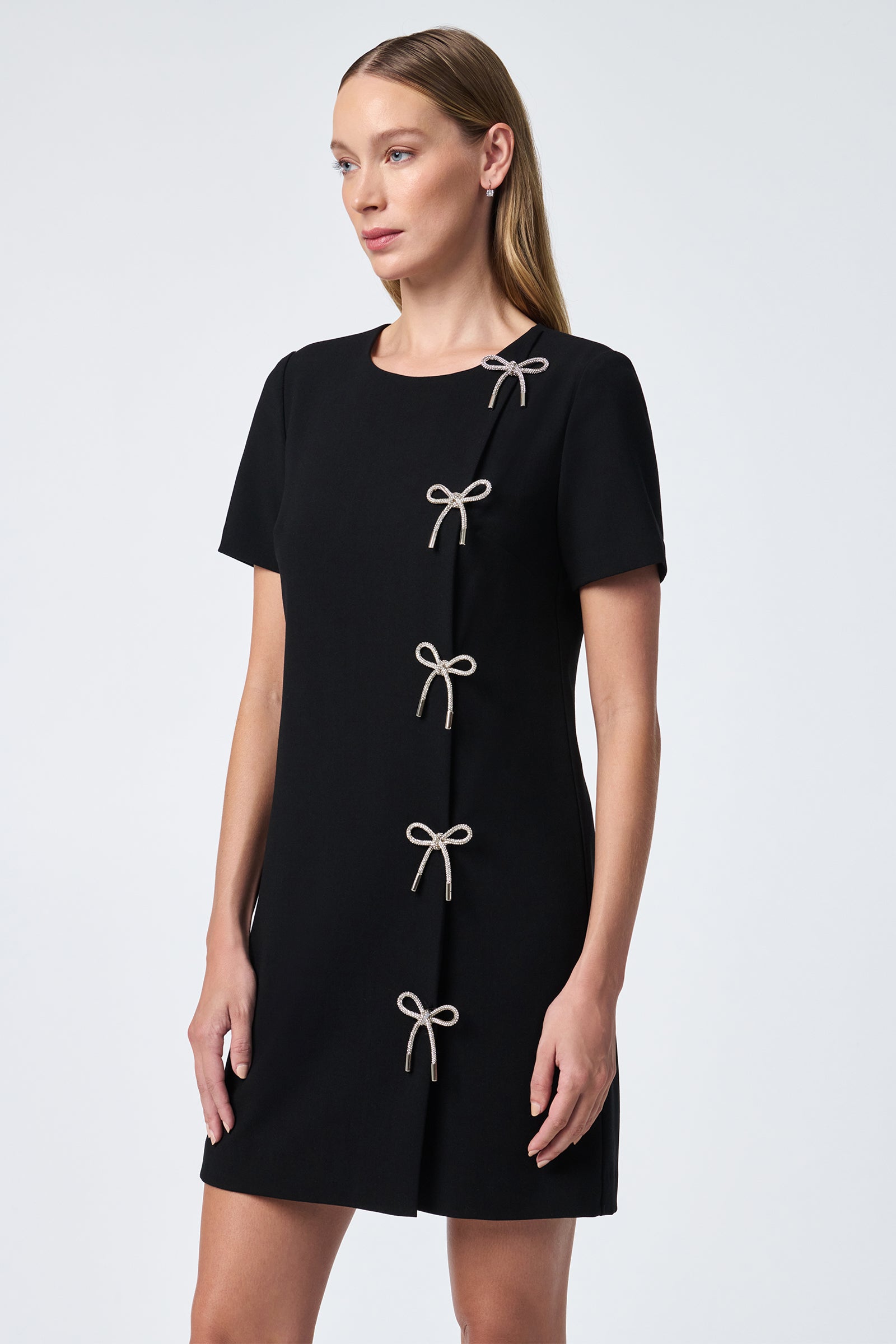 A woman wears the Diamonds Crystal Bows Sheath Dress—a short-sleeved, black sheath with five light bow accents diagonally from left shoulder to right hem.