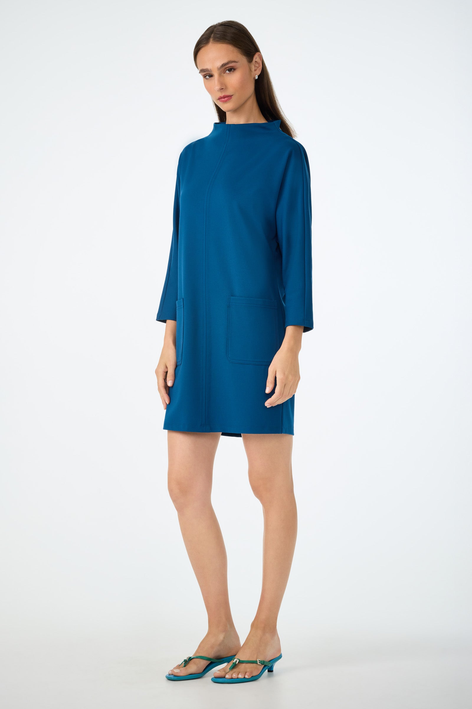 A woman with long brown hair and a neutral expression stands against a white background, wearing the blue Caracas Mini Dress—loose-fitting, knee-length, with long sleeves and two front pockets—paired with teal sandals.