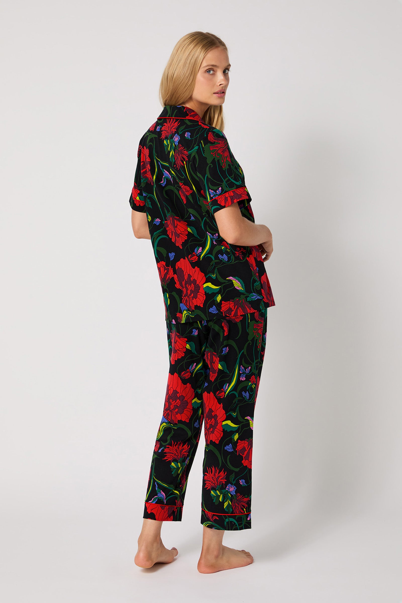 A woman with long blonde hair stands barefoot, slightly turned away, wearing the Haute Floral S/S Cropped PJ Set—black with bold red, green, and blue floral patterns—against a plain white background.