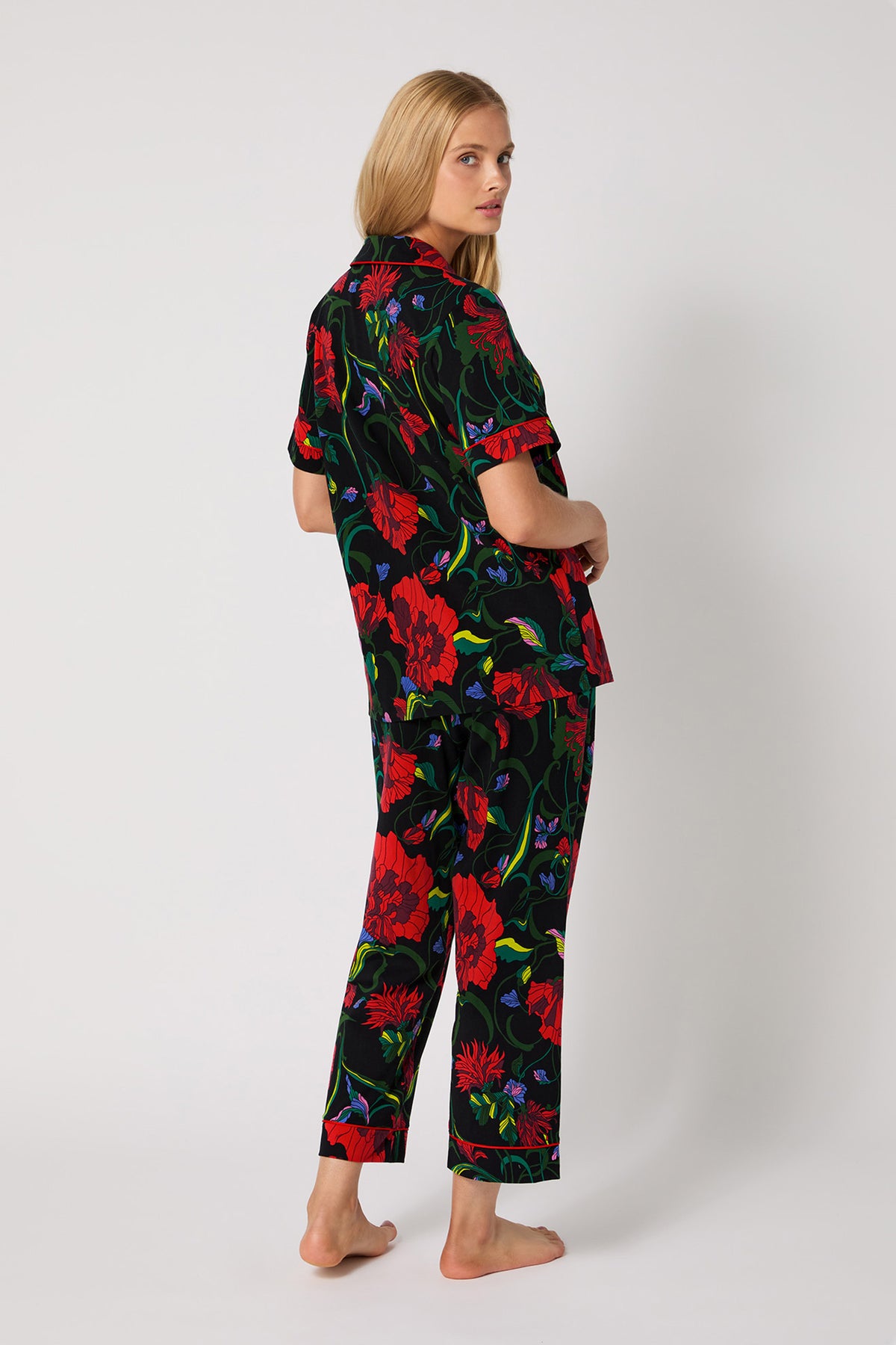 A woman with long blonde hair stands barefoot, slightly turned away, wearing the Haute Floral S/S Cropped PJ Set—black with bold red, green, and blue floral patterns—against a plain white background.