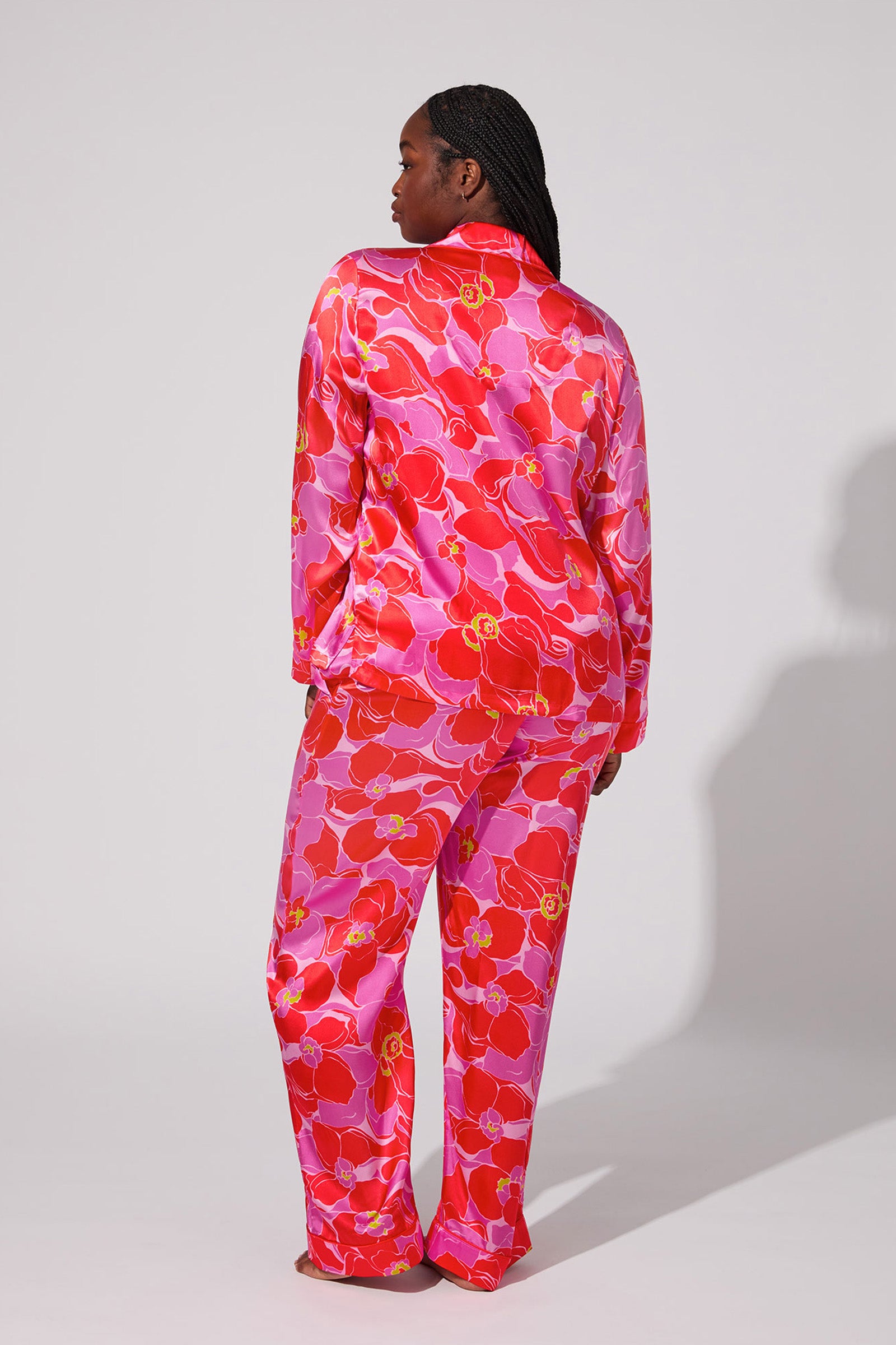 A person stands with their back to the camera, wearing the Silk Classic PJ Set - Winter Orchids, which features a striking red, pink, and yellow floral print. The backdrop is plain and light-colored.
