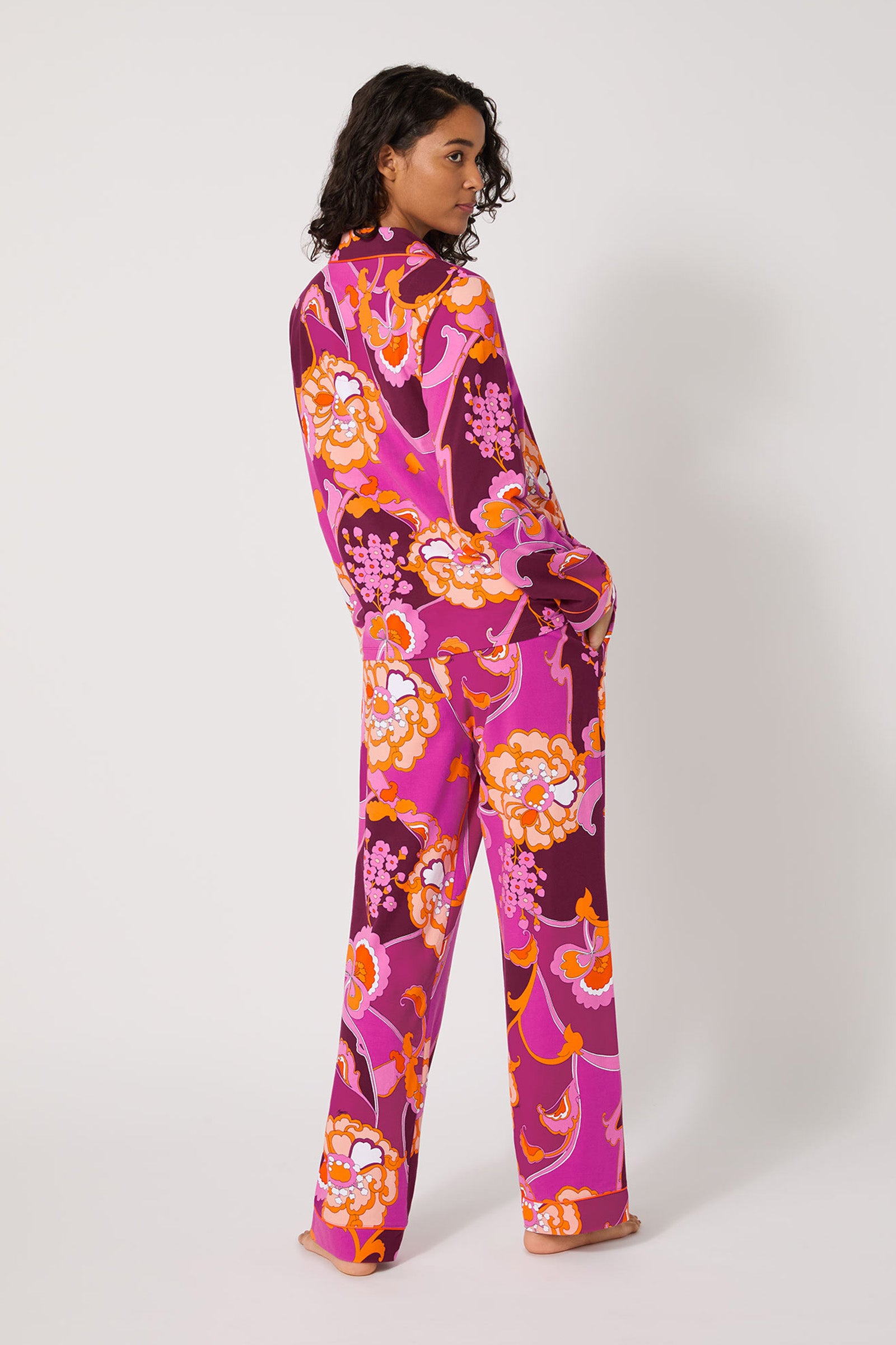 A person stands barefoot, facing away and glancing over their shoulder, wearing the Apache Plume L/S Classic Pj, featuring a bold pink, orange, and white floral pattern on a white background.