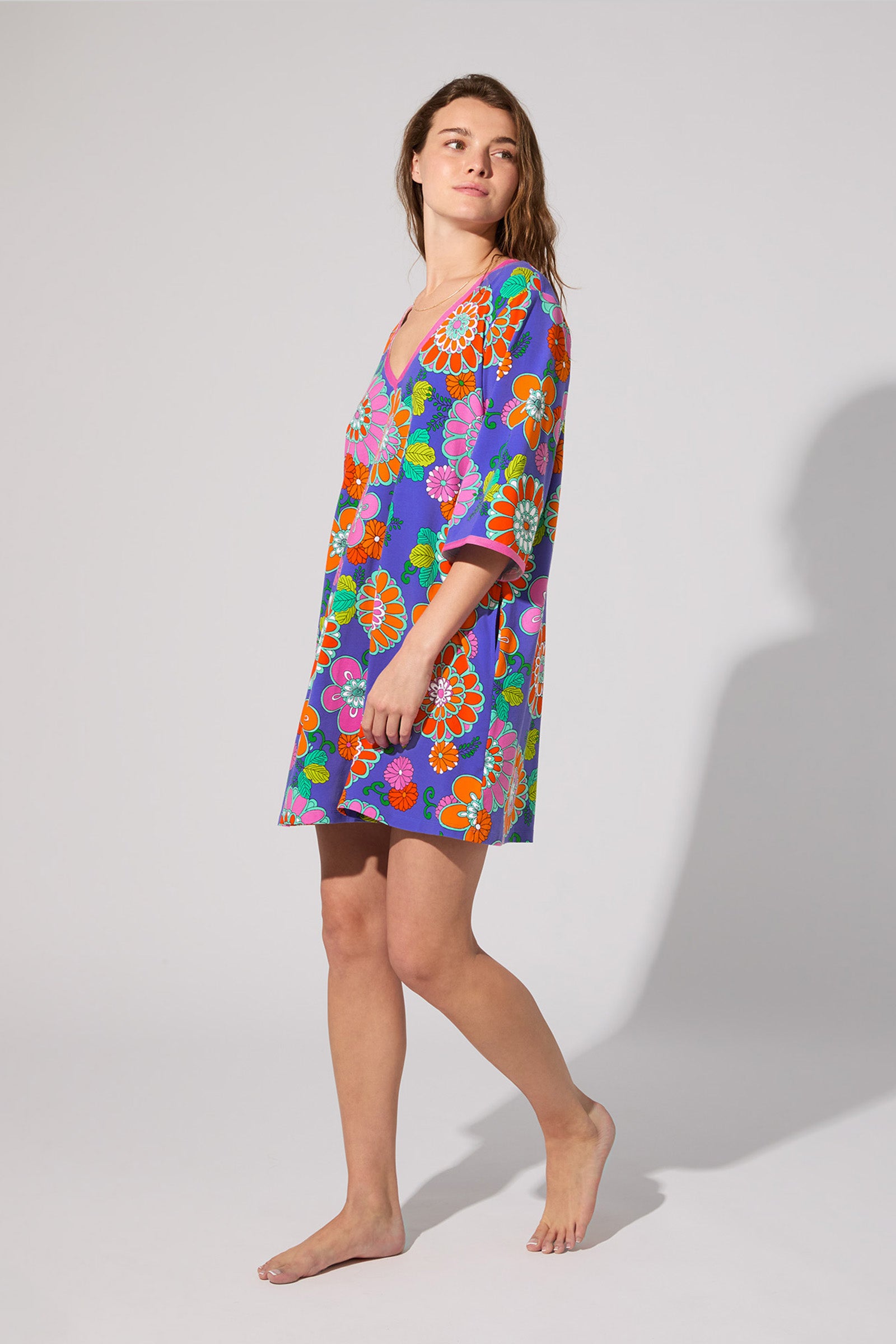 A woman stands barefoot in the PJ Caftan Dress - Tokyo Floral, featuring a vibrant floral print with a purple base, red, orange, green, and blue flowers. She is turned slightly sideways against a plain light gray background.