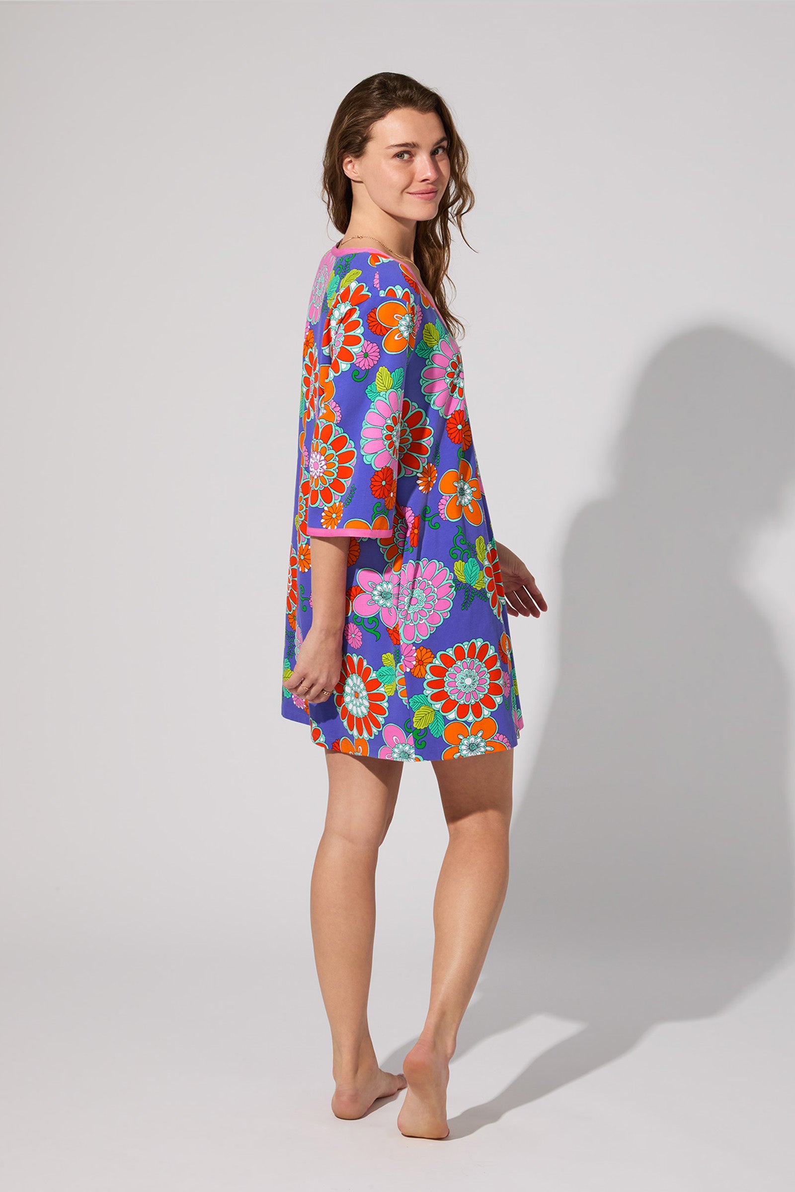 A woman stands barefoot, slightly turned, smiling gently in the PJ Caftan Dress - Tokyo Floral, a loose dress with red, orange, and white flowers on a blue background, against a plain light gray backdrop.