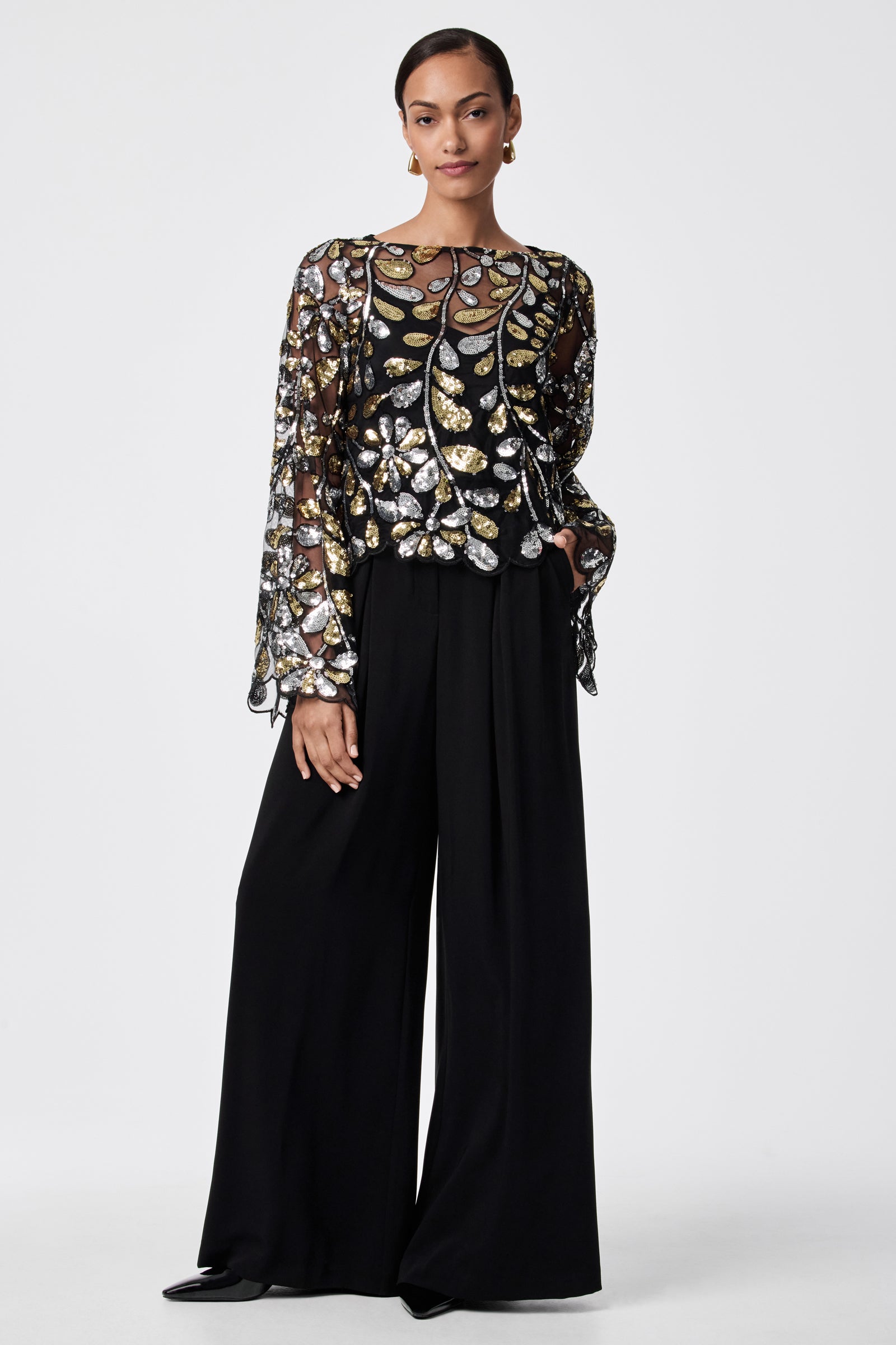 A woman with her hair pulled back and one hand in her pocket wears the FEMMES TOP—a long-sleeve, sheer black blouse made of 100% nylon with gold and silver sequin leaf patterns—paired with wide-leg black trousers.