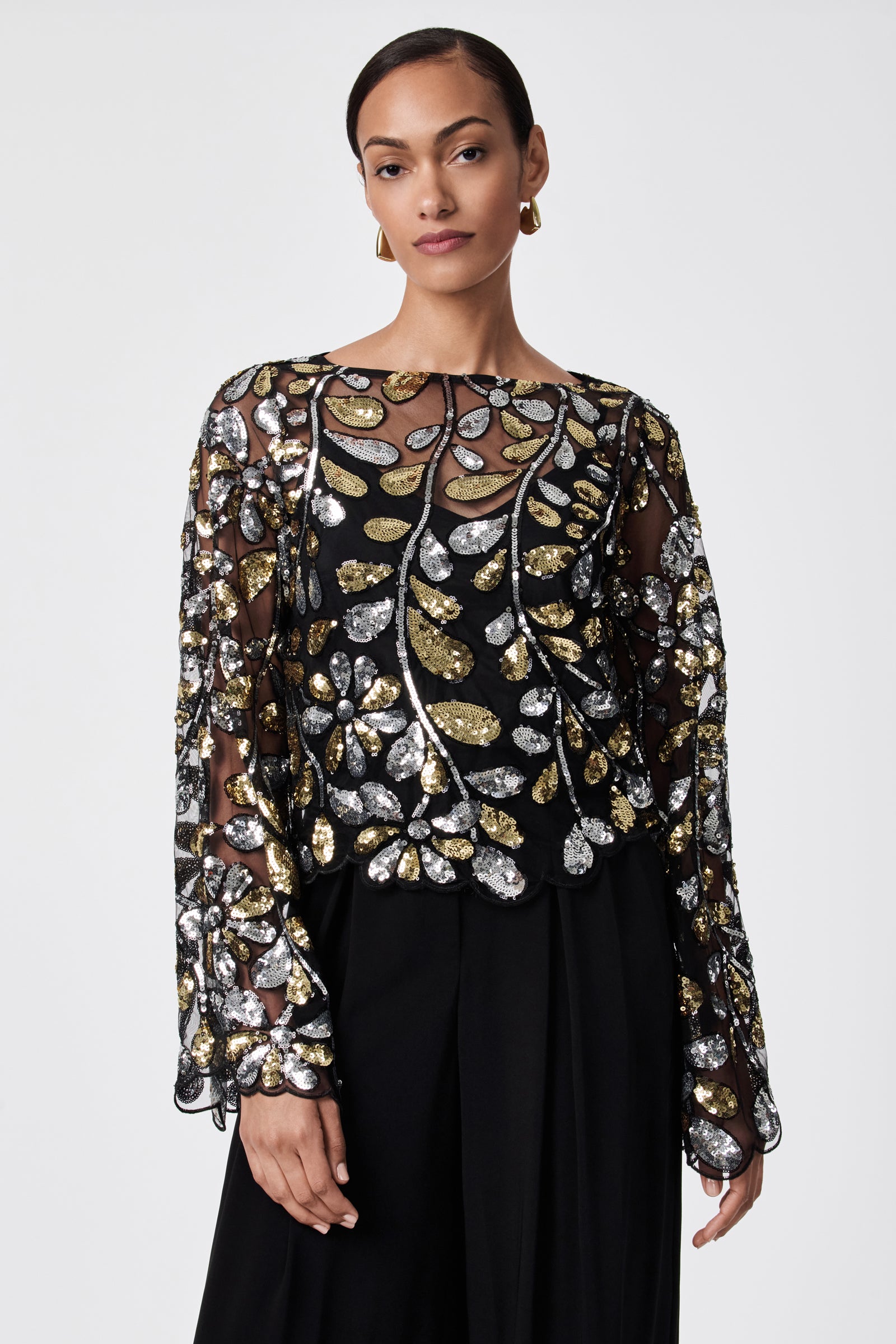 A woman with short hair and gold hoop earrings models the FEMMES TOP—a sheer black 100% nylon top with gold and silver sequin leaf patterns, long sleeves—paired with high-waisted black pants against a plain white background.