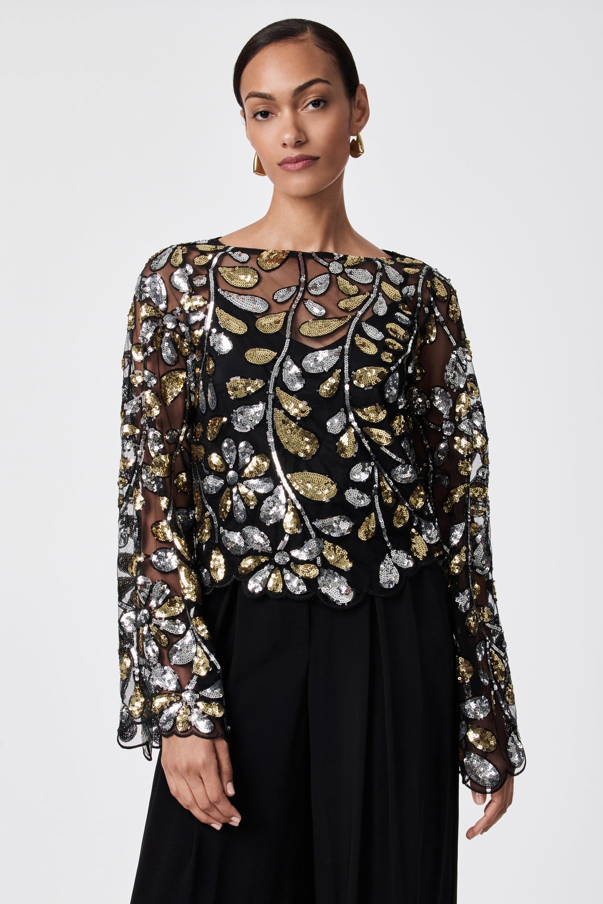 A woman with short hair and gold hoop earrings models the FEMMES TOP—a sheer black 100% nylon top with gold and silver sequin leaf patterns, long sleeves—paired with high-waisted black pants against a plain white background.