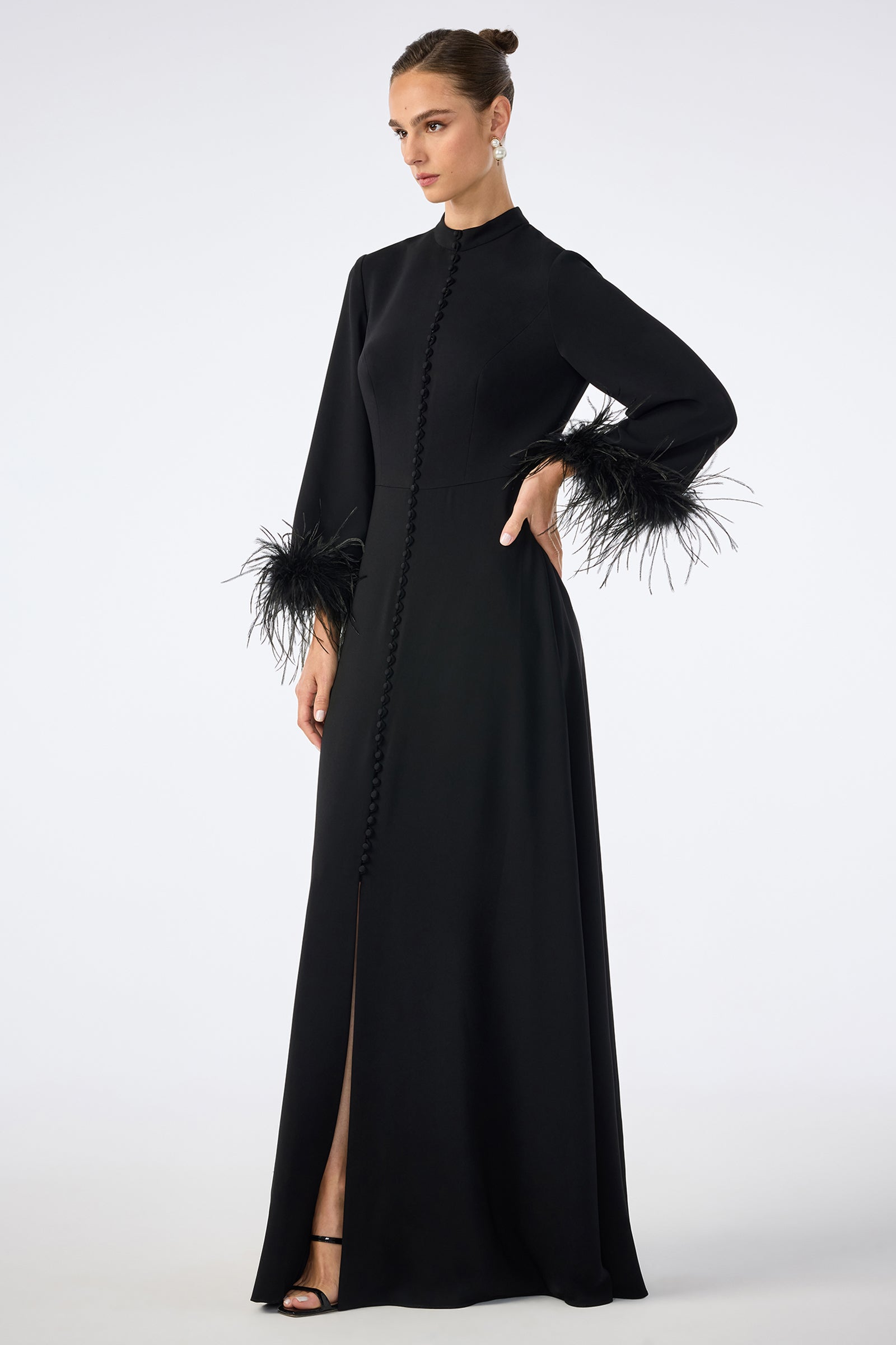 A woman wearing the Montmarte Dress—a long black gown with a high neckline, front buttons, and feathered cuffs—stands with one hand on her hip against a plain light background.
