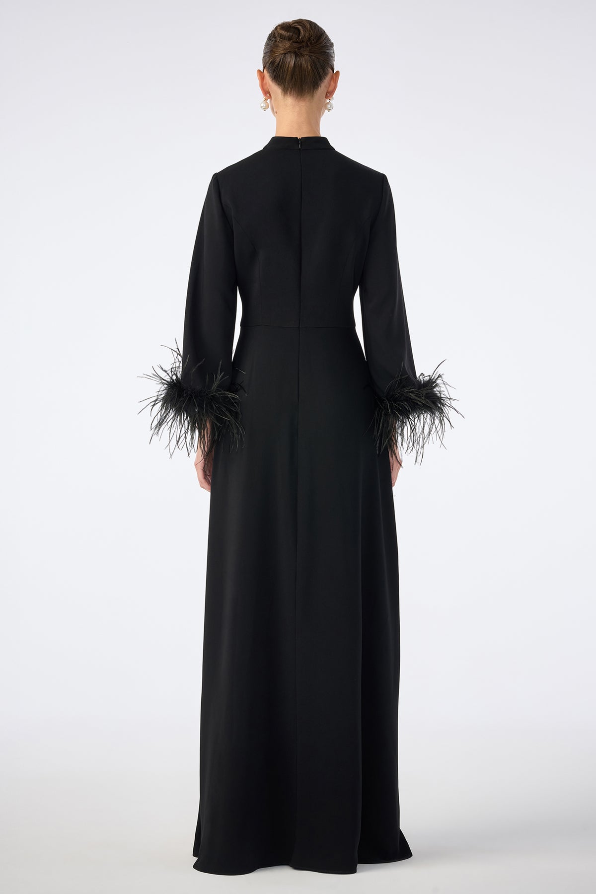 A person is shown from the back wearing the Montmarte Dress, a long black gown with long sleeves and feathered cuffs, standing against a plain white background.