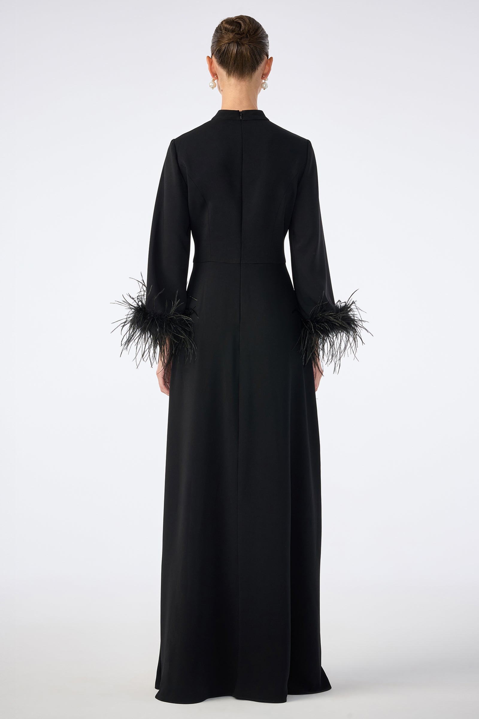 A person is shown from the back wearing the Montmarte Dress, a long black gown with long sleeves and feathered cuffs, standing against a plain white background.