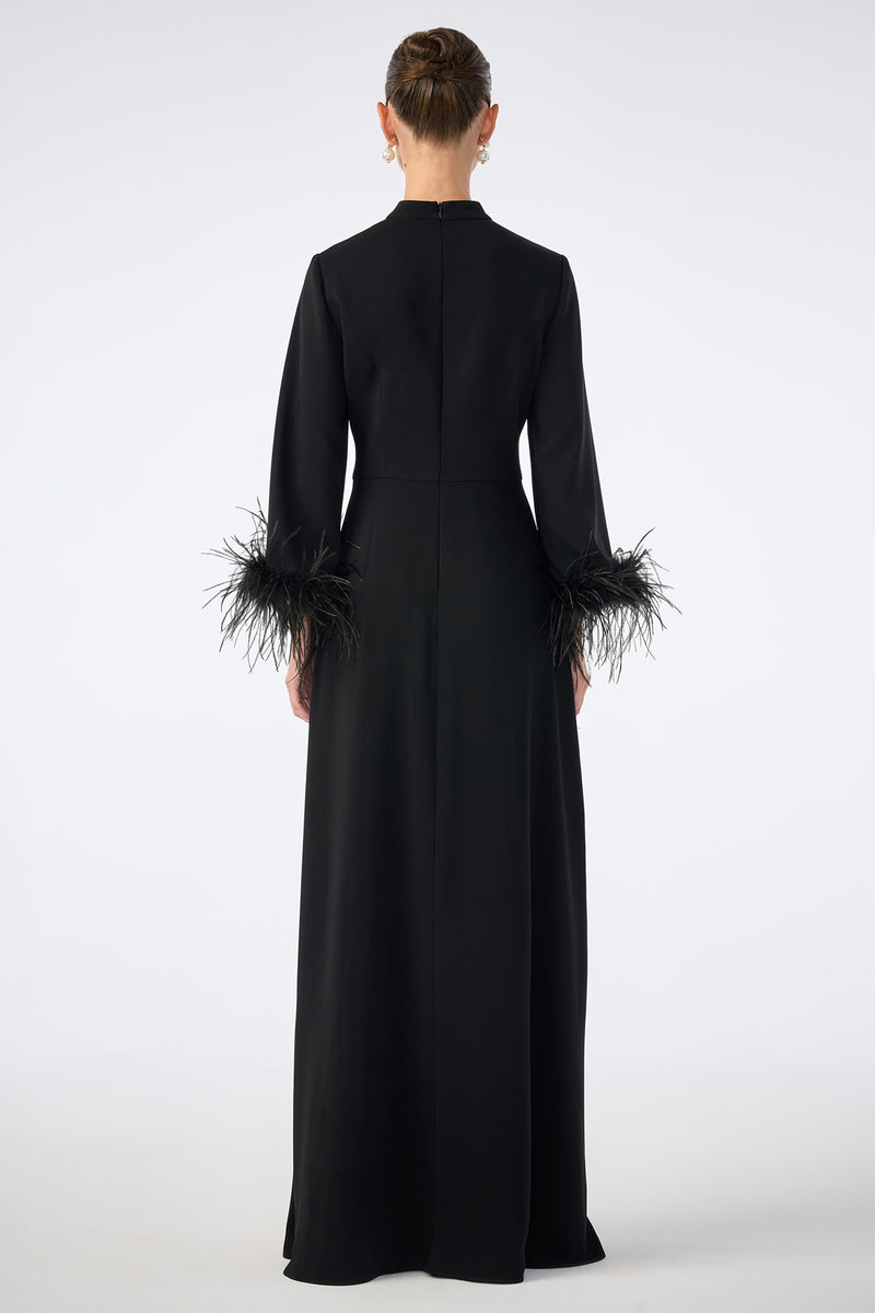 A person is shown from the back wearing the Montmarte Dress, a long black gown with long sleeves and feathered cuffs, standing against a plain white background.