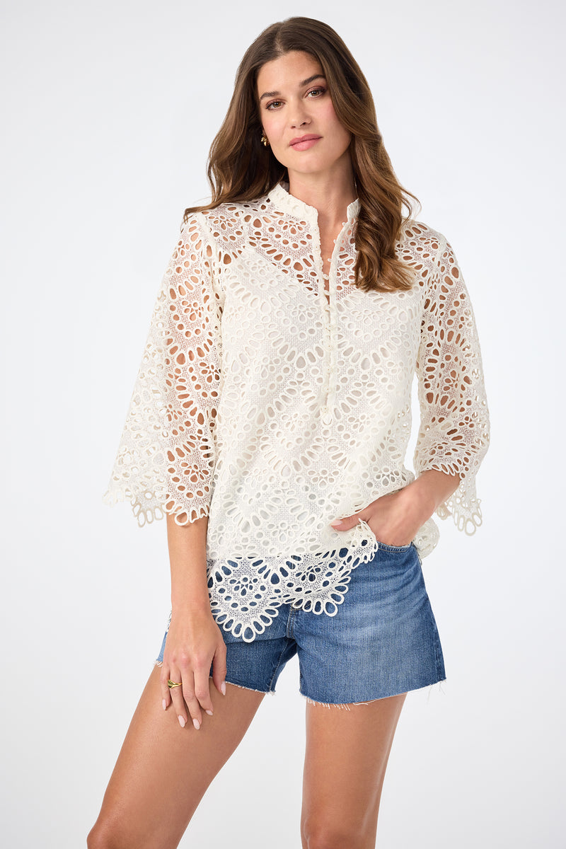A woman wears the Martigues Lace Top in Whitewash with blue denim shorts, standing against a plain light background and looking at the camera with a soft smile.