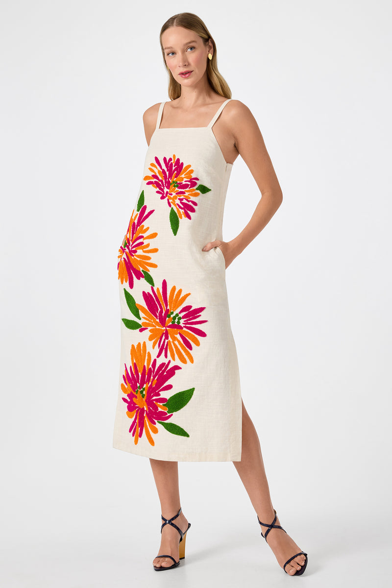 A woman wears the Yona Embroidered Flowers Midi Dress, a sleeveless, light-hued midi with bold pink, orange, and yellow floral prints. She has one hand in her pocket and pairs it with dark strappy heels.