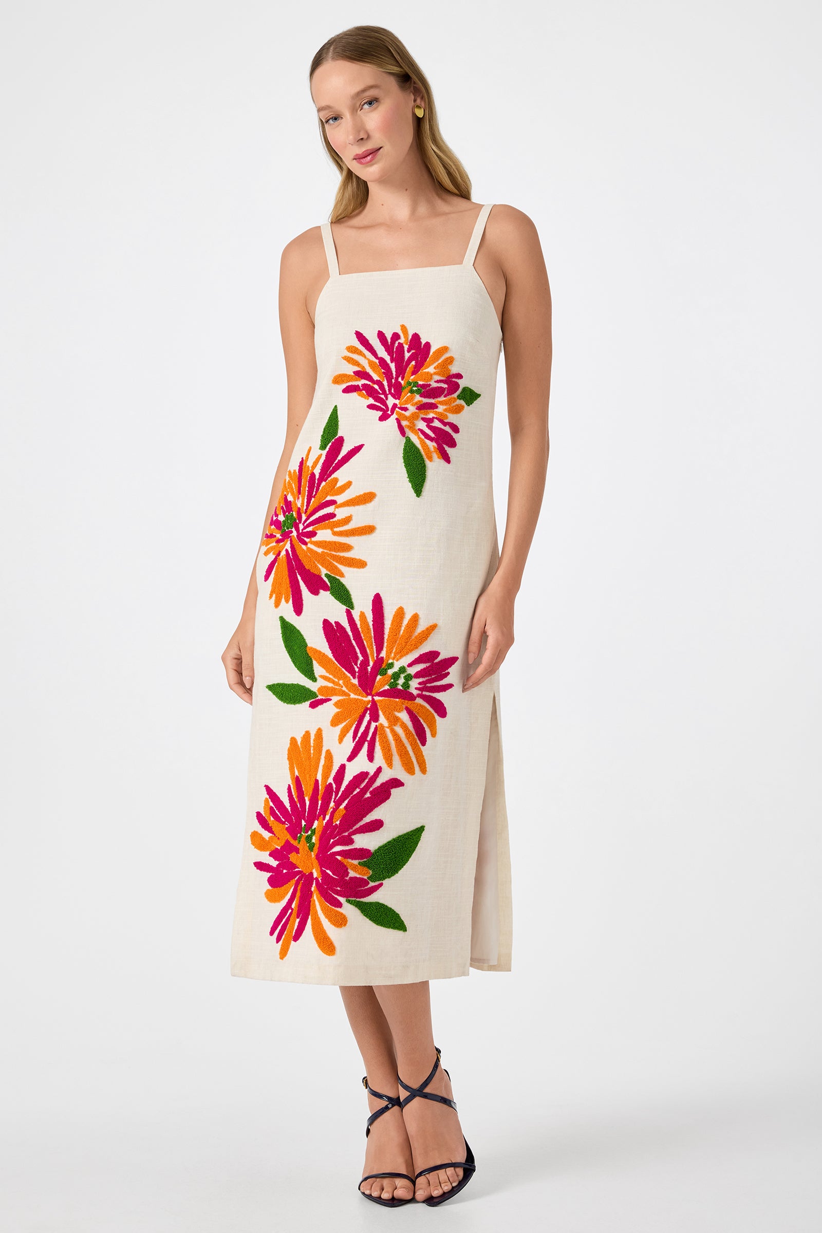 A woman wears the Yona Embroidered Flowers Midi Dress in Riku Embroidery, a cream sleeveless, ankle-length dress with wide straps and bold pink, orange, and green floral designs. She stands against a white background in black strappy sandals.