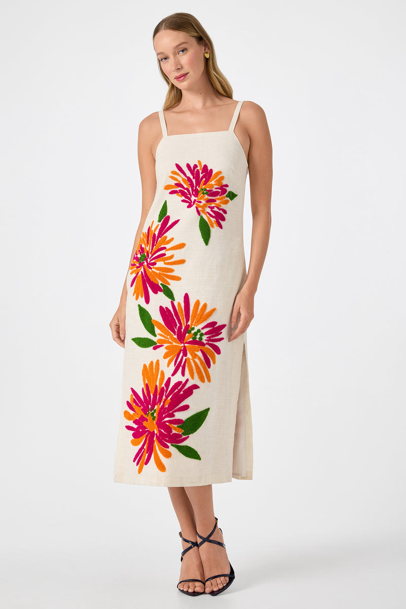 A woman wears the Yona Embroidered Flowers Midi Dress in Riku Embroidery, a cream sleeveless, ankle-length dress with wide straps and bold pink, orange, and green floral designs. She stands against a white background in black strappy sandals.
