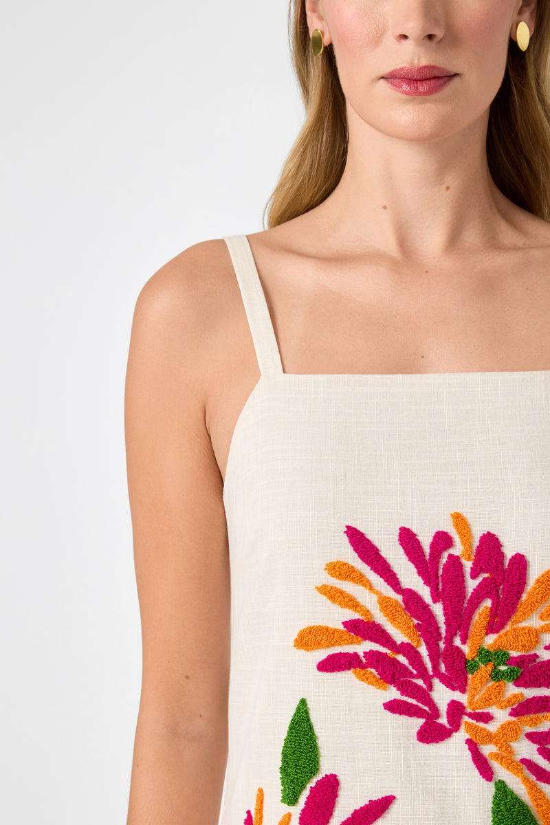 A woman wears the Yona Embroidered Flowers Midi Dress, a sleeveless beige dress with bright pink, orange, and green floral embroidery, gold round earrings, and her blonde hair down, shown from the shoulders up against a white background.