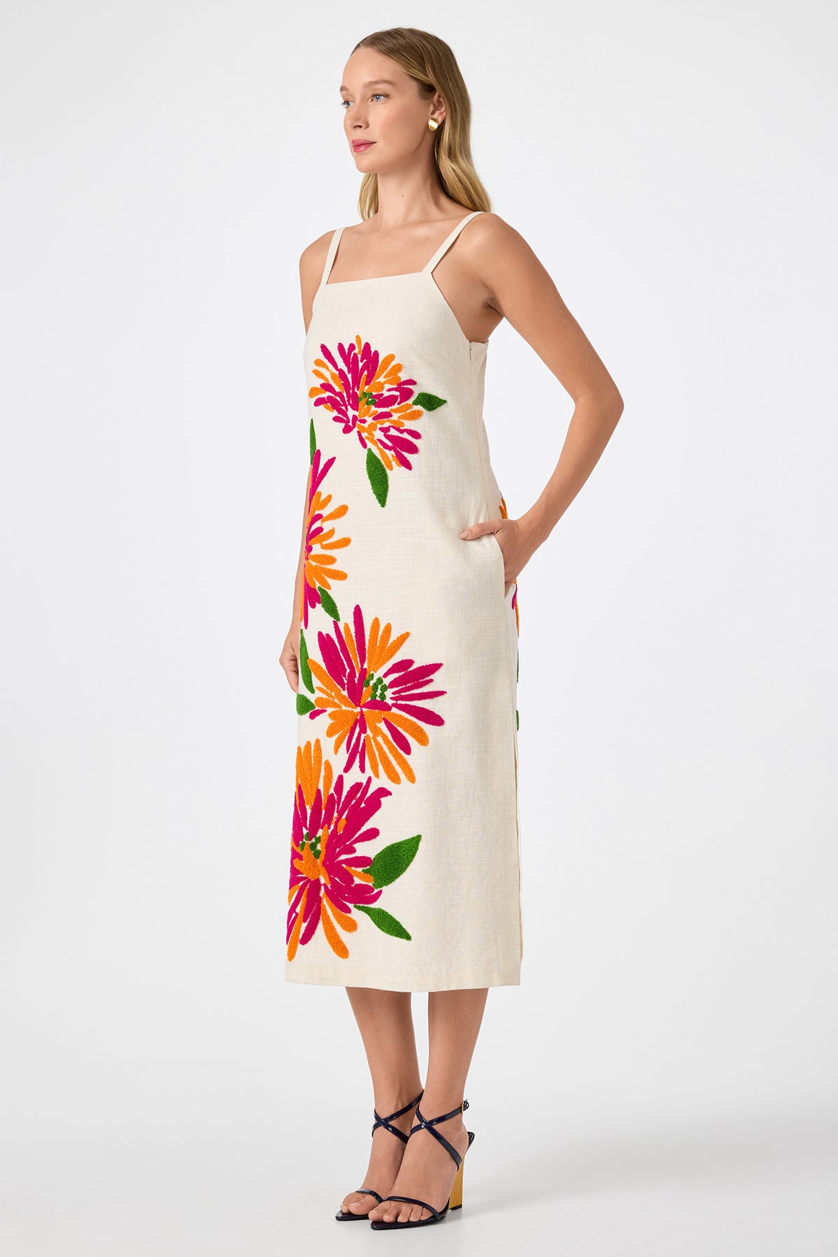 A woman poses in the Yona Embroidered Flowers Midi Dress, a sleeveless, natural-toned dress with thin straps, pink-orange-green floral embroidery, and pockets. She pairs it with strappy high-heeled sandals.