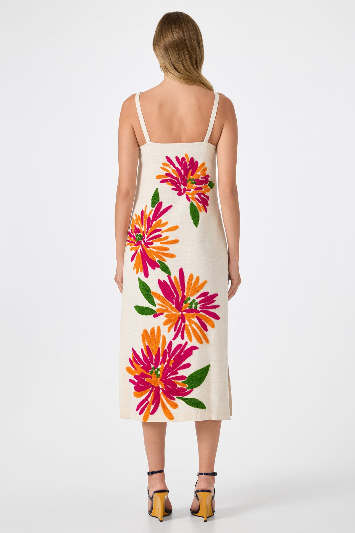A woman with light brown hair faces away from the camera, wearing the Yona Embroidered Flowers Midi Dress, a cream sleeveless midi adorned with large pink, orange, and green floral motifs. She pairs it with strappy heels.