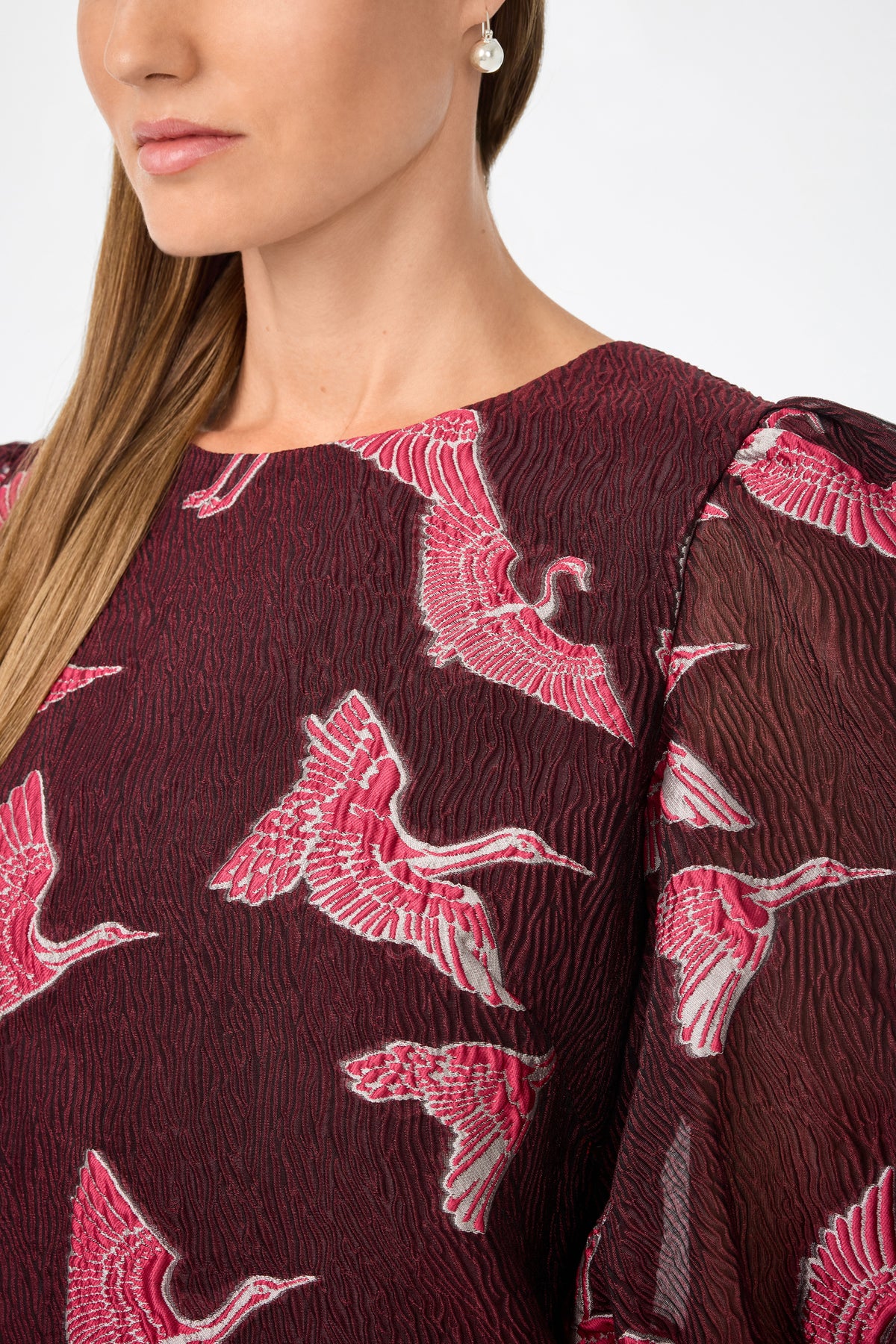 A woman models the Ringo Short Puff Sleeve Mini Dress—a textured burgundy piece with pink and white crane embroidery. Only her lower face, a single earring, and part of her straight hair are visible.