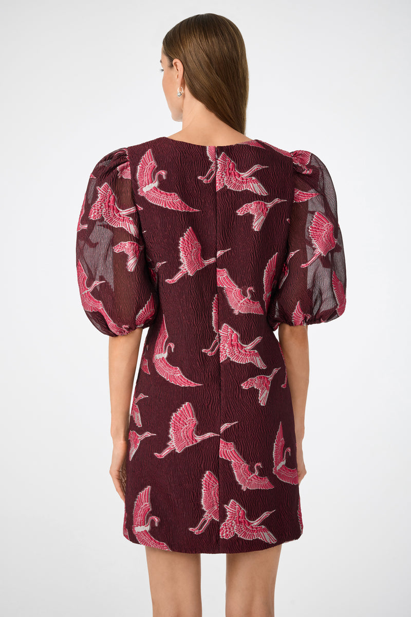 A woman with straight brown hair models the Ringo Short Puff Sleeve Mini Dress in maroon, which has textured fabric, pink and white crane patterns, and puffed mid-length sleeves. She is shown from the back against a plain background.