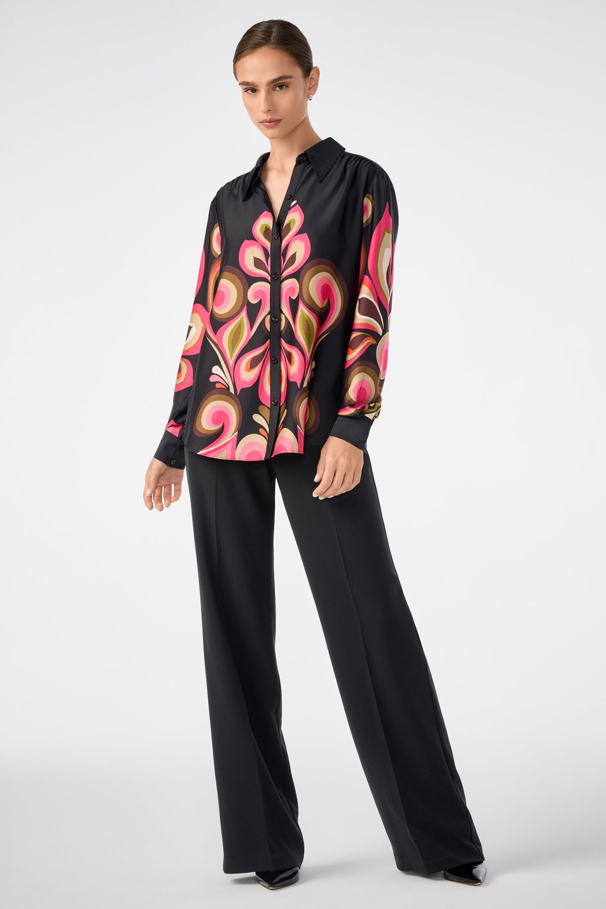A woman wears the Cassavetes Border Shirt by Sanctuary, featuring a bold pink and beige abstract print, paired with black wide-leg trousers and pointed shoes against a plain light backdrop.