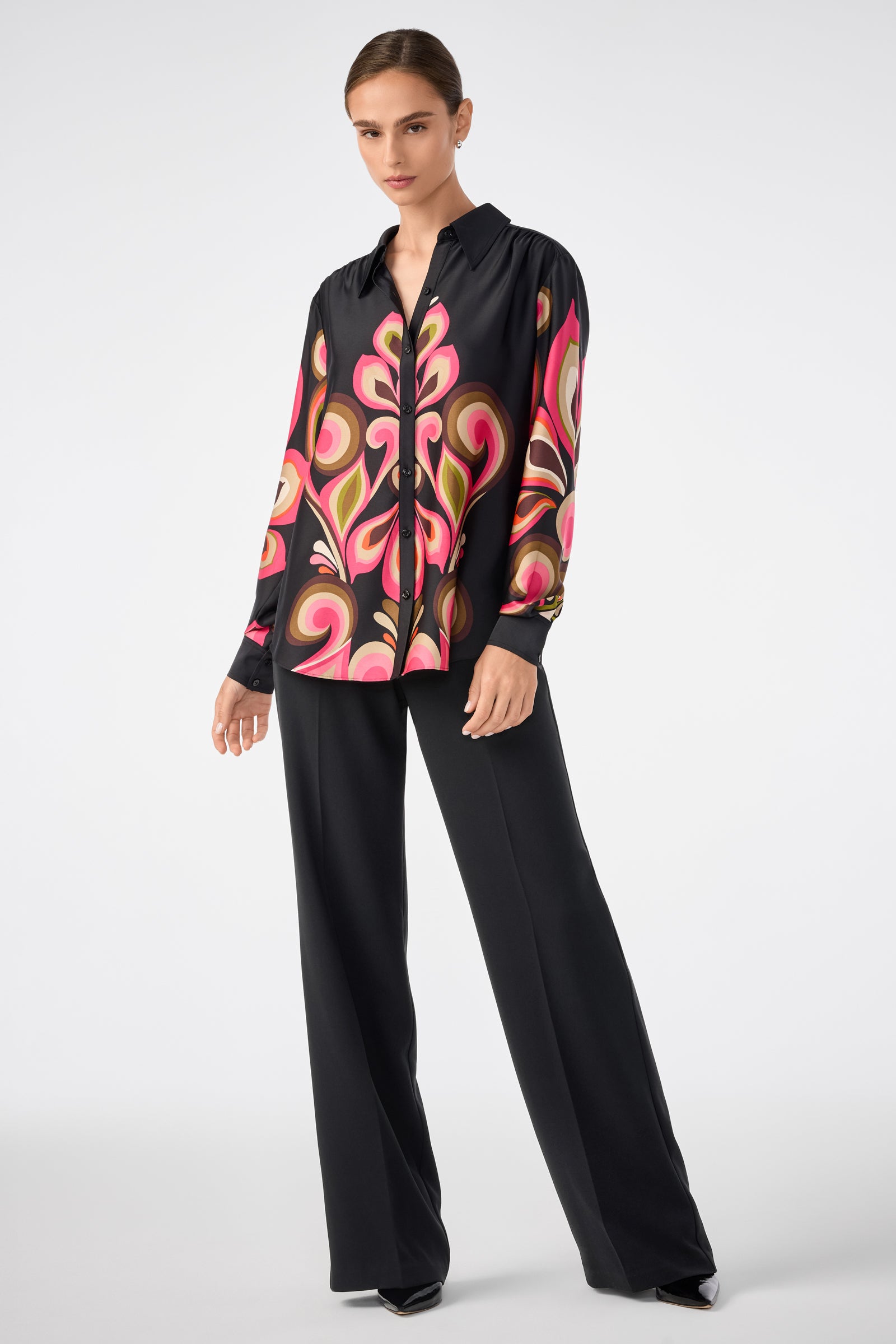 A woman wears the Cassavetes Border Shirt by Sanctuary, featuring a bold pink and beige abstract print, paired with black wide-leg trousers and pointed shoes against a plain light backdrop.