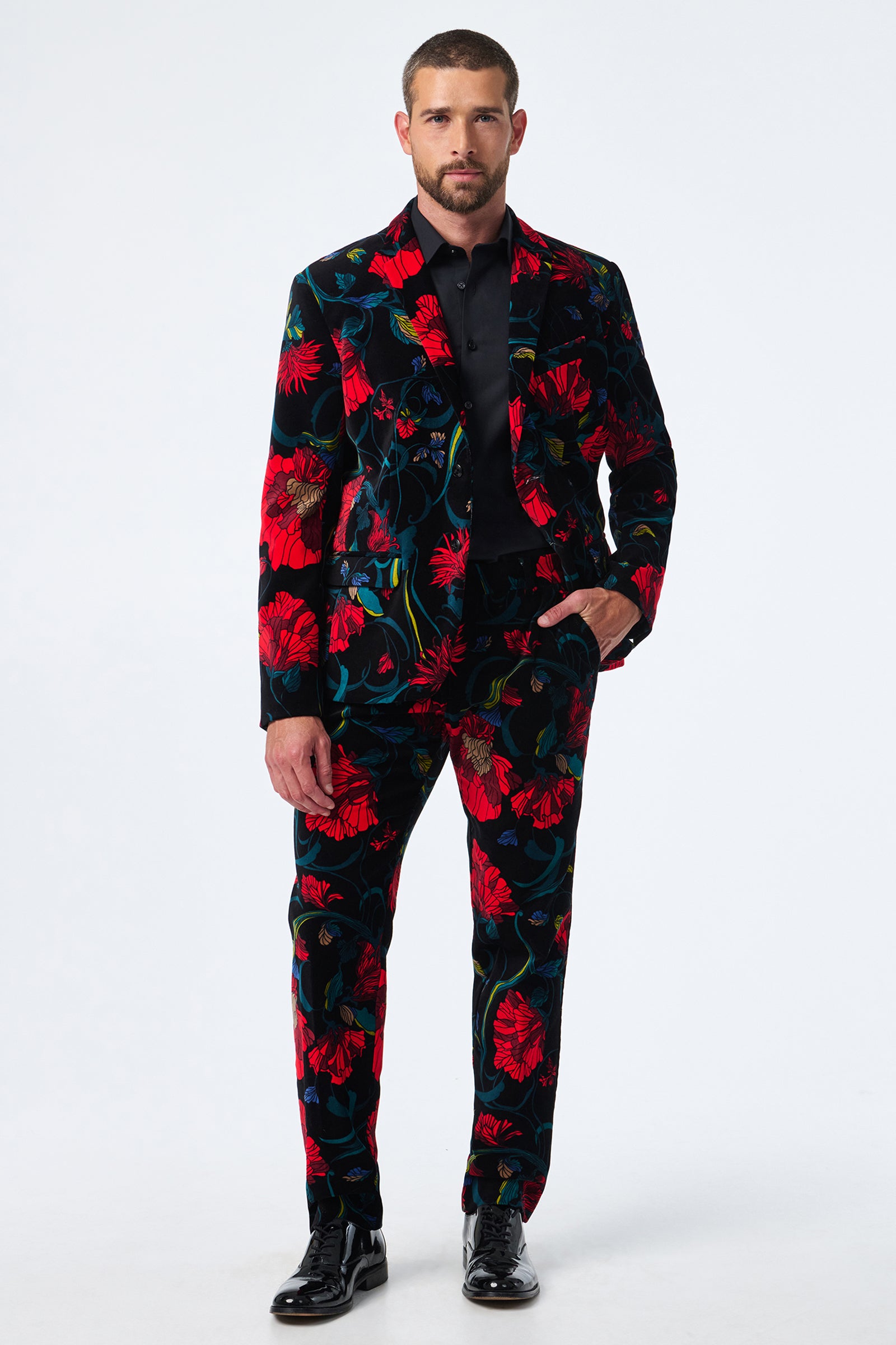 A man stands against a plain background wearing the Thurston Printed Velvet Blazer, featuring a bold red and green floral pattern, paired with a black shirt and shiny black dress shoes.