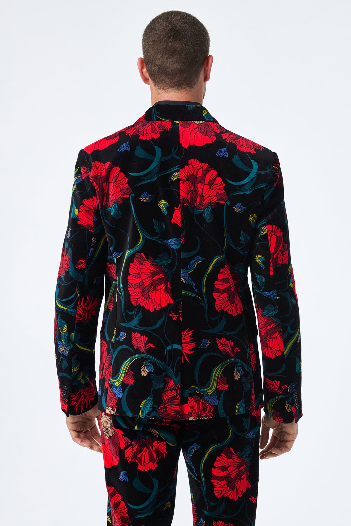 A man is seen from behind in the Thurston Printed Velvet Blazer, featuring a black base with vibrant red floral patterns and green and blue accents; he has short hair and stands with relaxed arms at his sides.