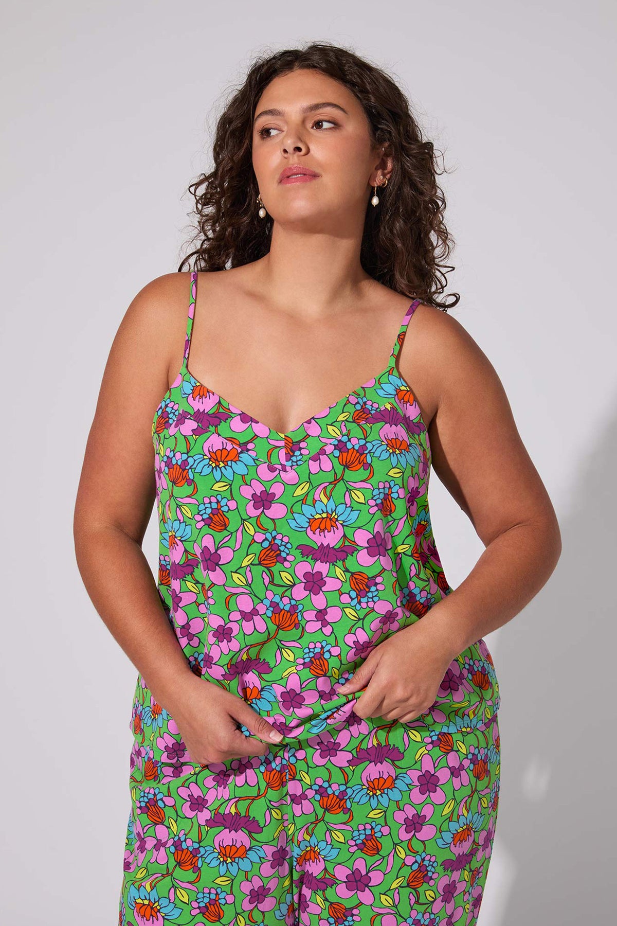 A woman with curly brown hair wears the Cami PJ Set - Garden Floral, featuring a colorful floral pattern and thin straps, as she stands against a plain light background looking slightly to the side.