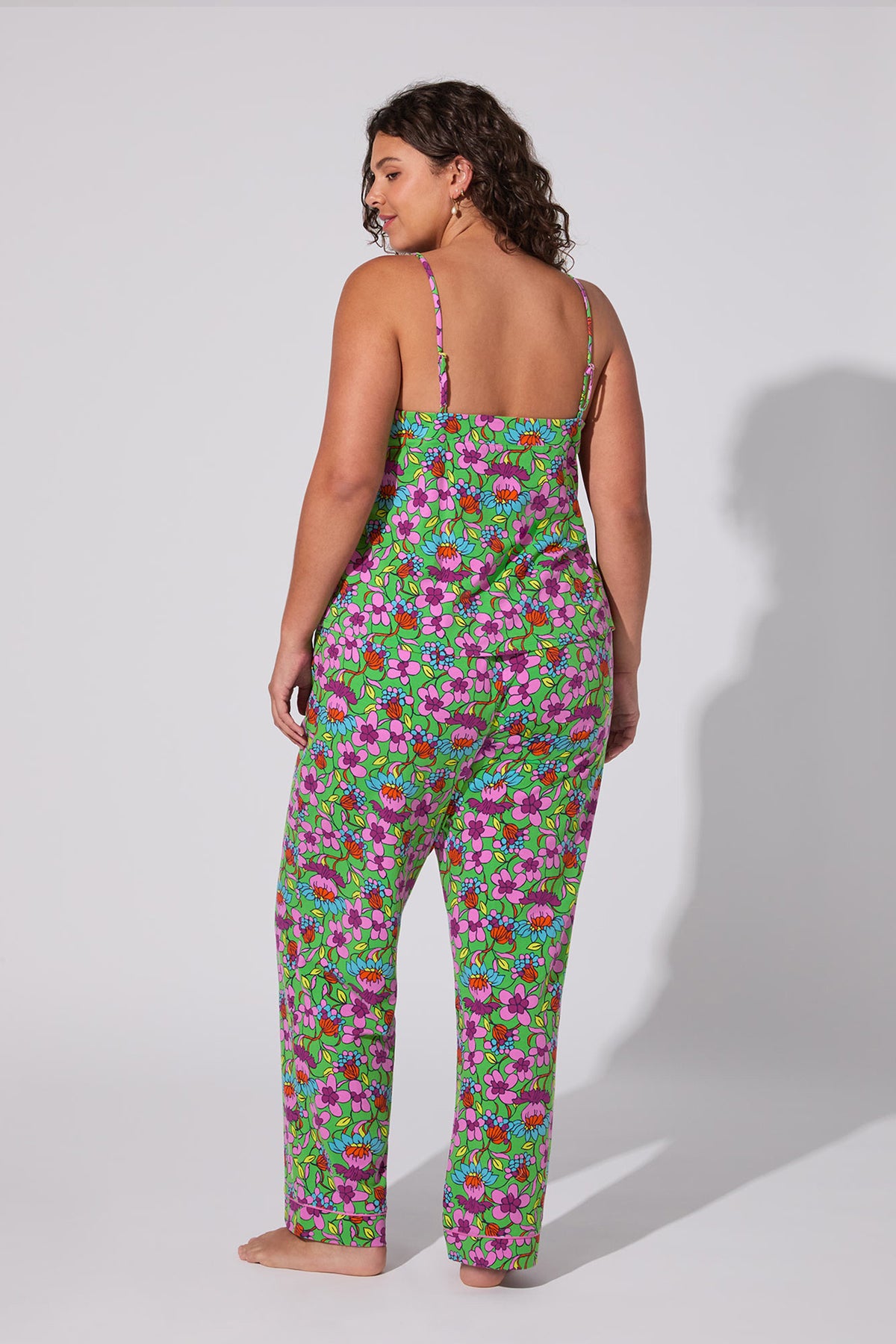 A woman with curly hair stands barefoot, facing away from the camera, wearing the Cami PJ Set - Garden Floral, a colorful floral sleepwear piece with pink, green, and purple patterns against a light gray background.