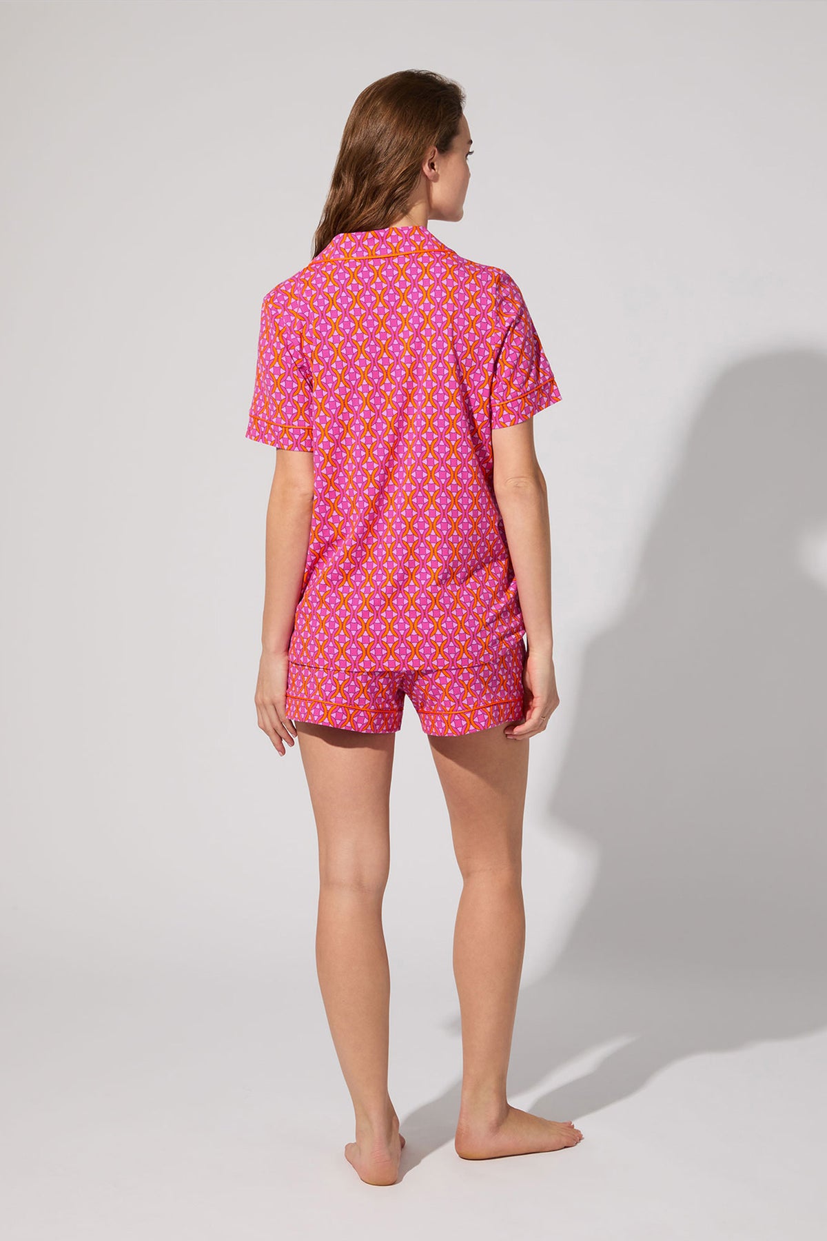 A woman stands barefoot facing away, wearing the Shorty PJ Set - Chain Geo, a matching pink short-sleeve pajama set with a yellow and white floral pattern, against a plain light background.