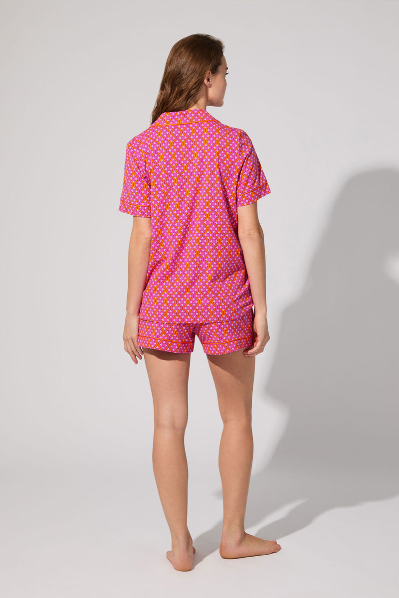 A woman stands barefoot facing away, wearing the Shorty PJ Set - Chain Geo, a matching pink short-sleeve pajama set with a yellow and white floral pattern, against a plain light background.