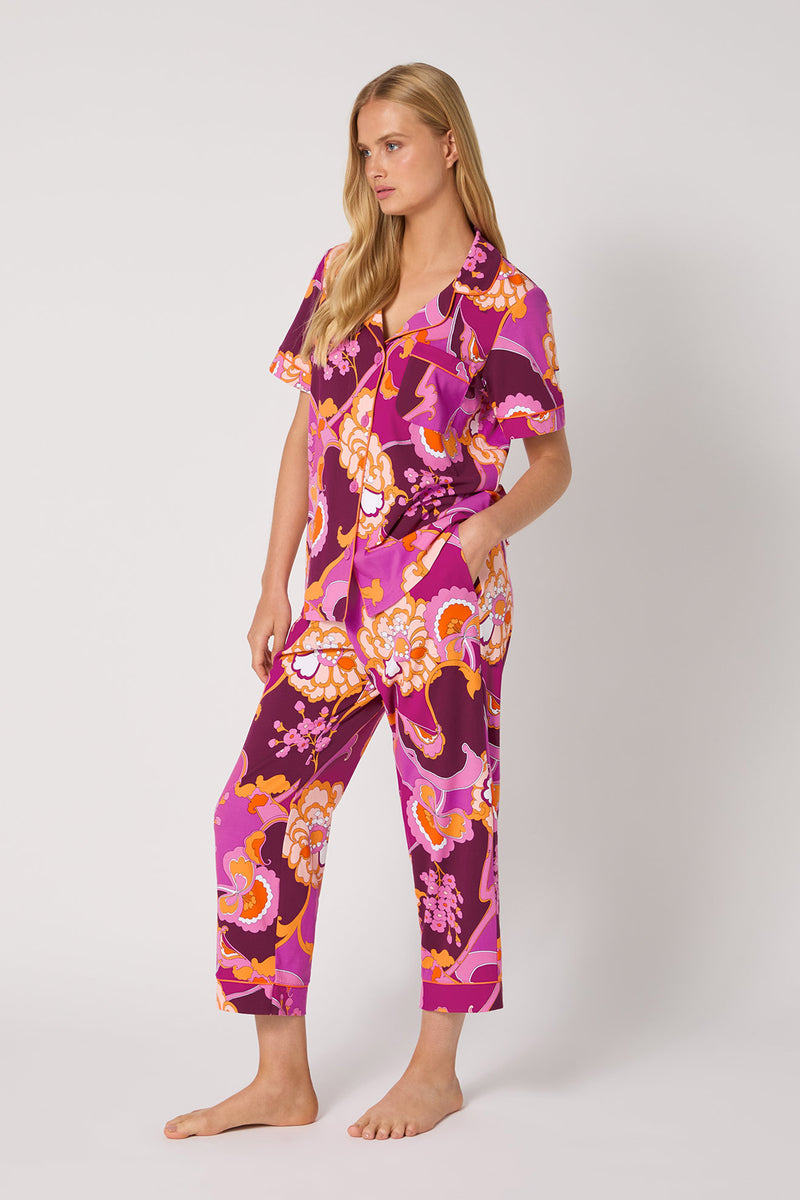 A woman stands barefoot in the Apache Plume S/S Cropped Pj, a matching set with vibrant pink, purple, and orange floral patterns, featuring short sleeves and cropped pants against a plain light background.