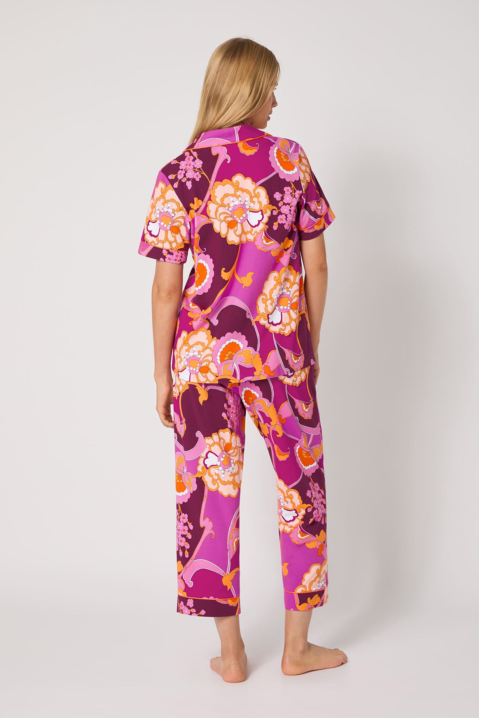 A person stands barefoot facing away, wearing the Apache Plume S/S Cropped Pj— a matching set featuring a vibrant pink, purple, orange, and white floral pattern on a light background.