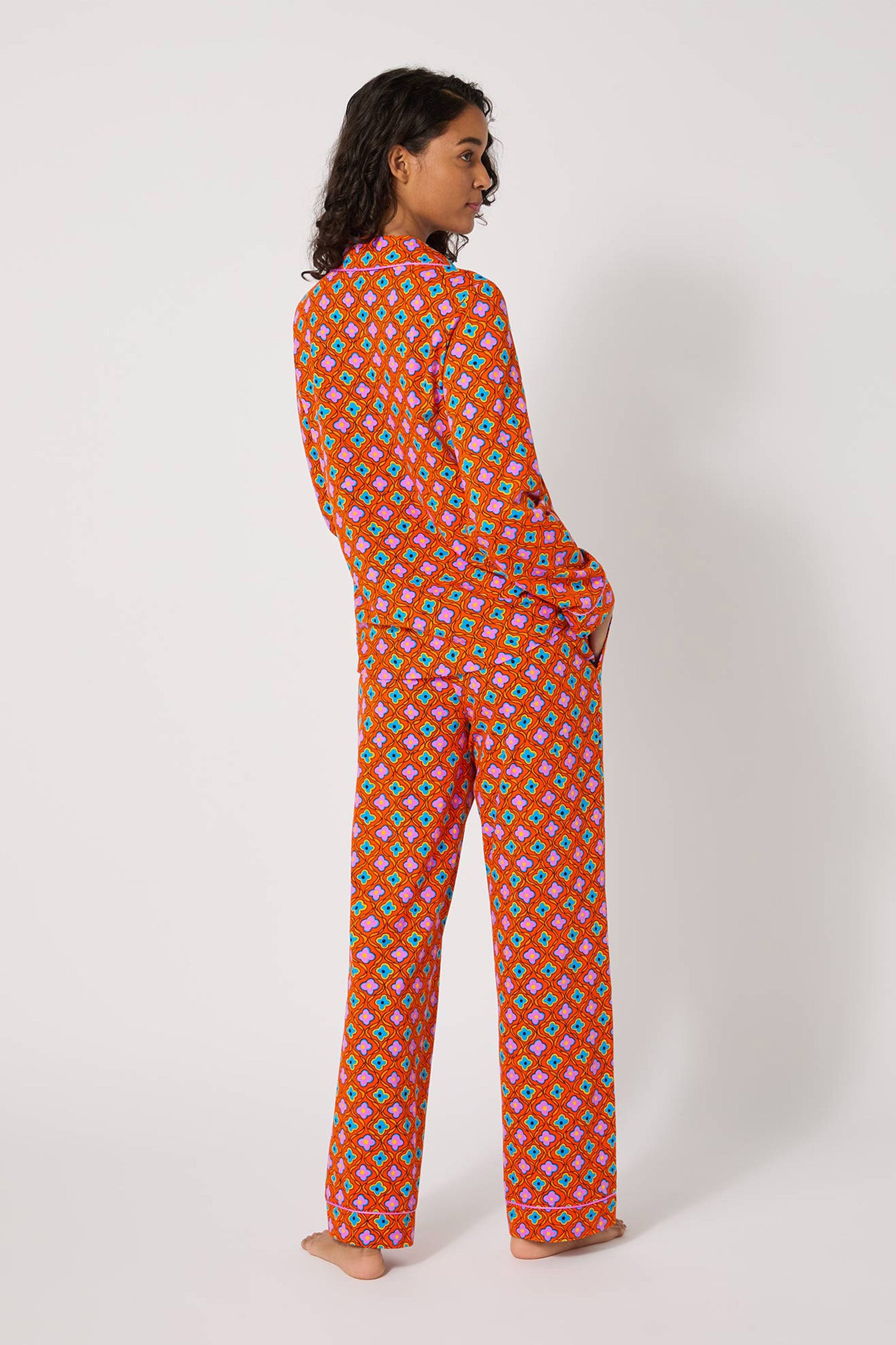 A person stands barefoot, facing slightly away from the camera, wearing the Daisy Foulard L/S Classic Pj—a matching red pajama set with a colorful geometric pattern. The background is plain white.