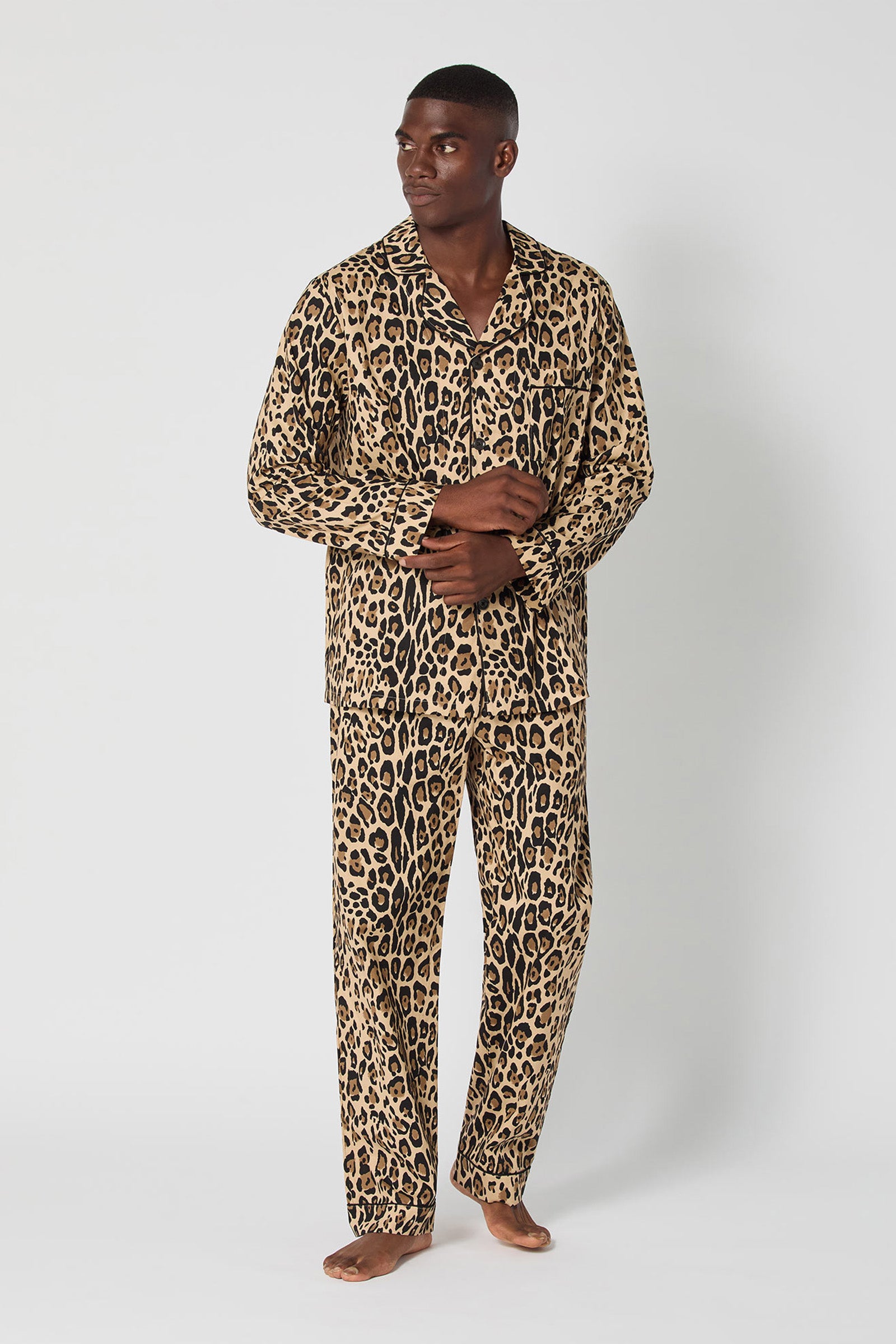 A man with short hair stands barefoot against a white background, wearing the Leopard Stripe Mens Classic Pj set featuring a button-up shirt and long pants in matching leopard print. He looks to the side with his hands loosely clasped.