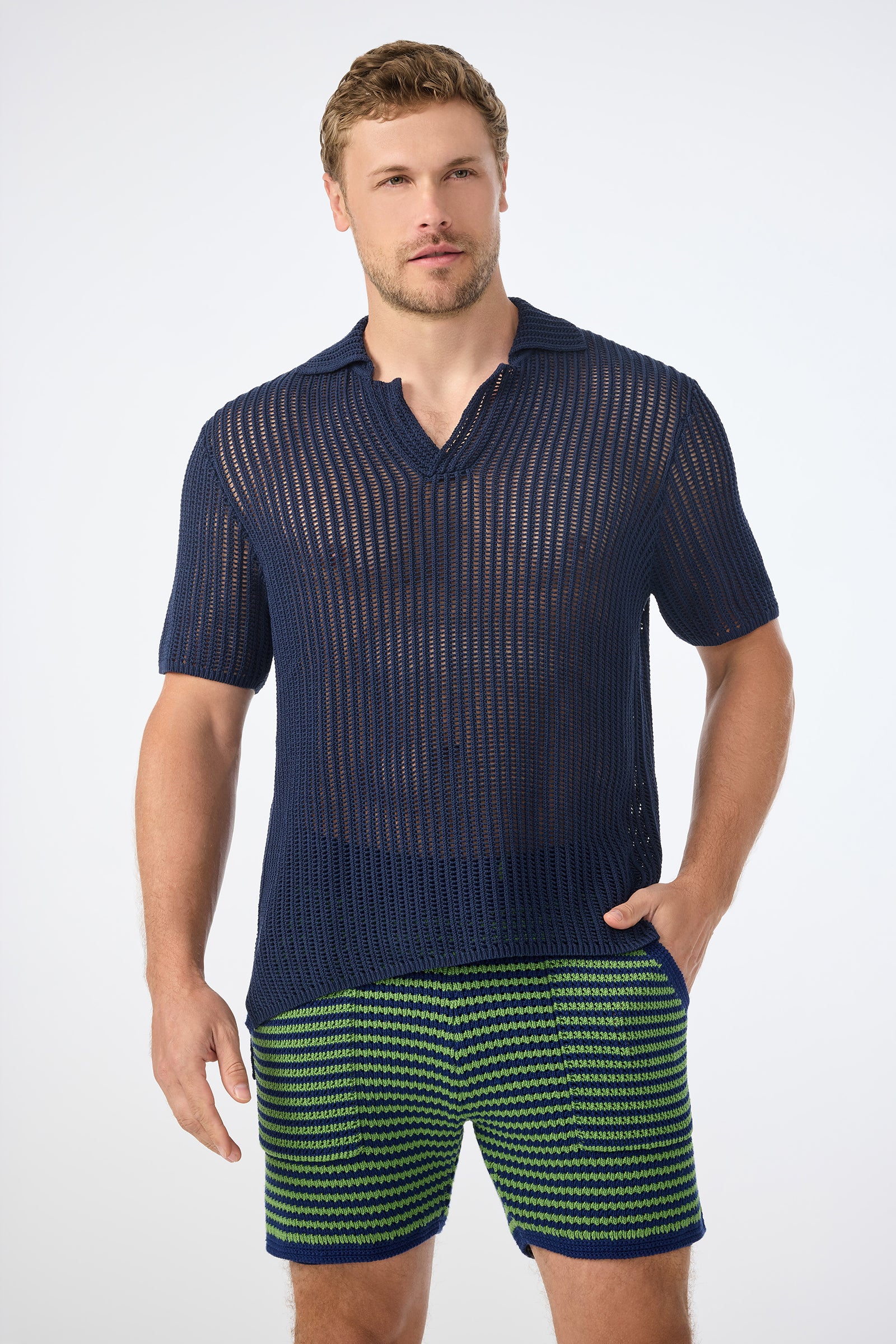 A man wearing the Javi Crochet Pullover in Noh Nightfall stands with one hand in his pocket, paired with green patterned shorts, against a plain white background.