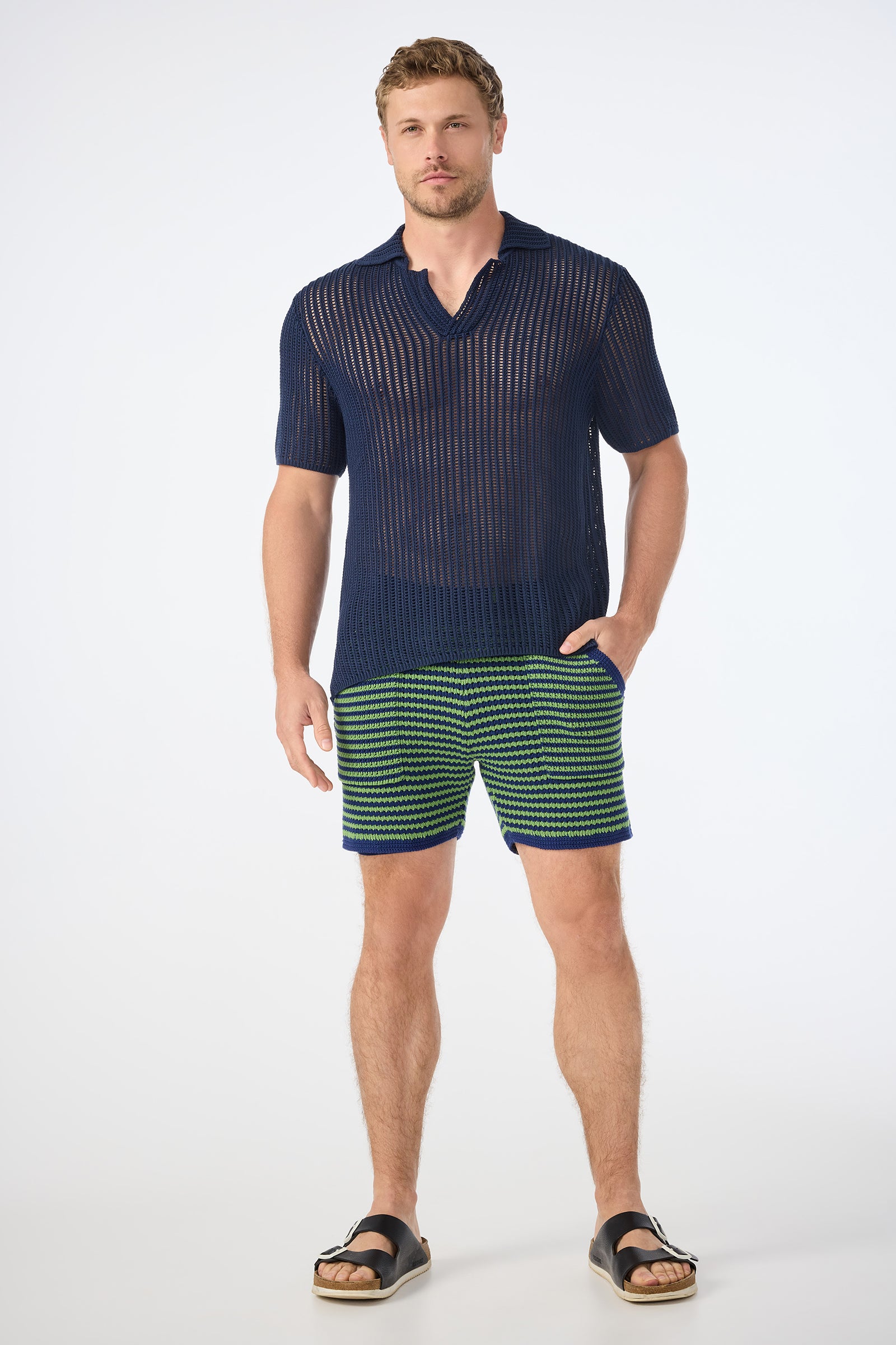A man poses against a plain background wearing the Javi Crochet Pullover - Noh Nightfall, paired with green and navy striped shorts and black sandals. He stands with one hand in his pocket and a neutral expression.