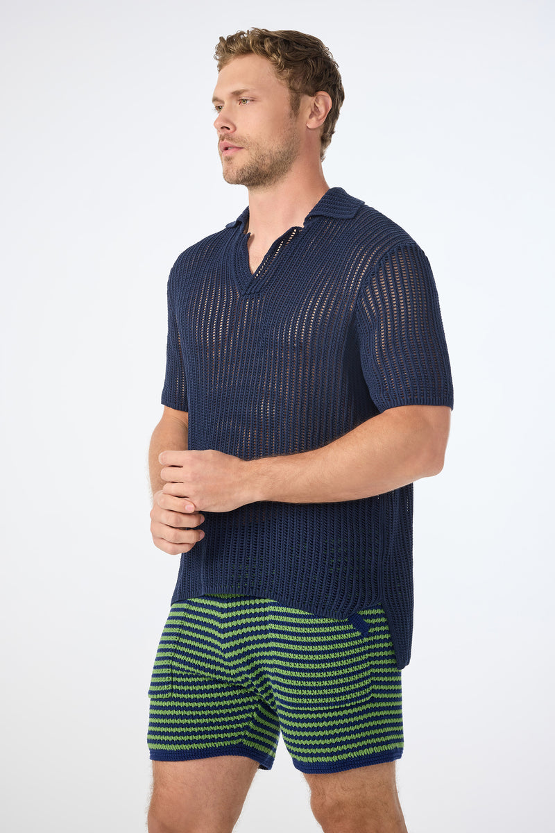 A man stands against a plain background wearing the Javi Crochet Pullover - Noh Nightfall, looking to his left with his hands lightly clasped.