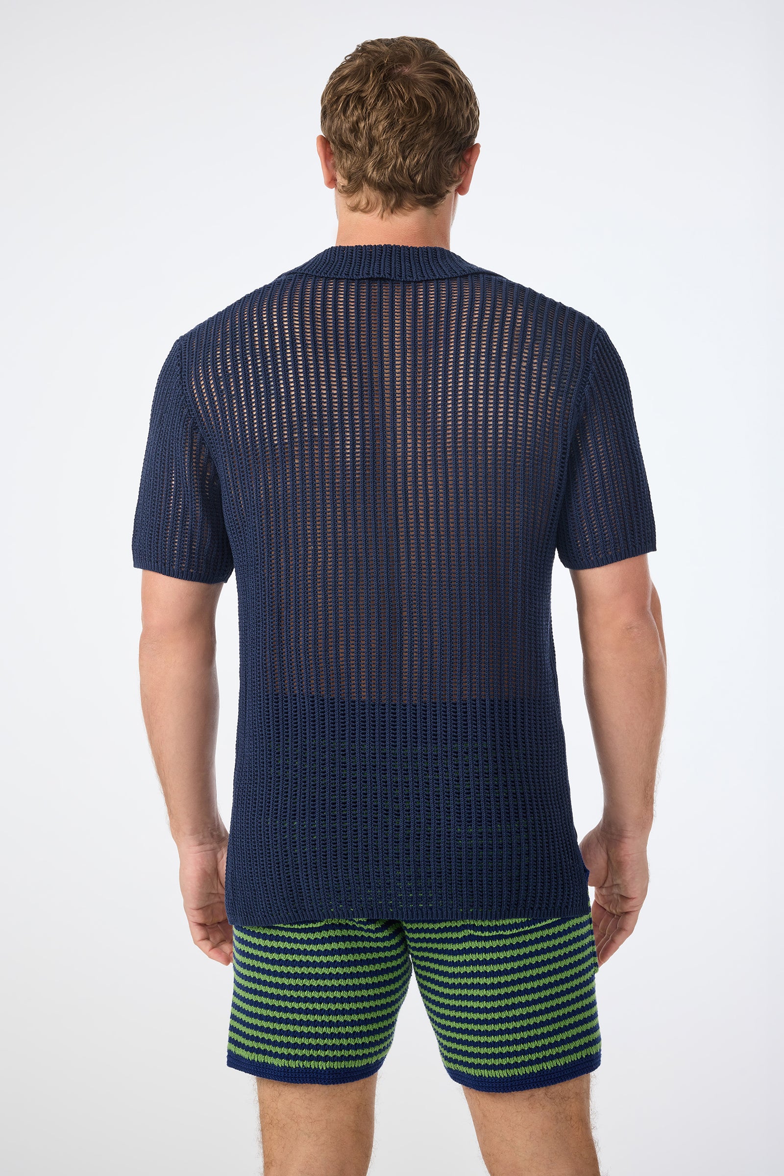 A man is seen from behind wearing the Javi Crochet Pullover - Noh Nightfall, a navy blue sheer knit short-sleeve shirt, paired with green-and-blue striped shorts against a plain light background.