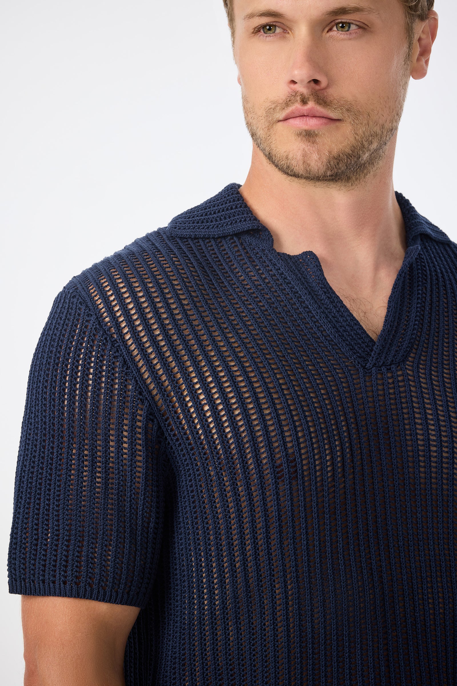 A man wears the Javi Crochet Pullover - Noh Nightfall, a dark blue, short-sleeve, open-collar knit shirt with a see-through ribbed design, shown from the chest up against a plain background.