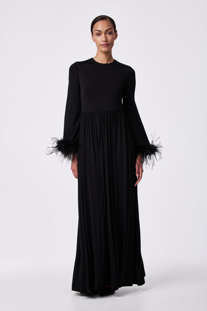 A woman wears the Orla Feather-Cuff Maxi Dress, featuring a fitted bodice, round neckline, long sleeves with black feather cuffs, and a floor-length skirt against a plain white background.