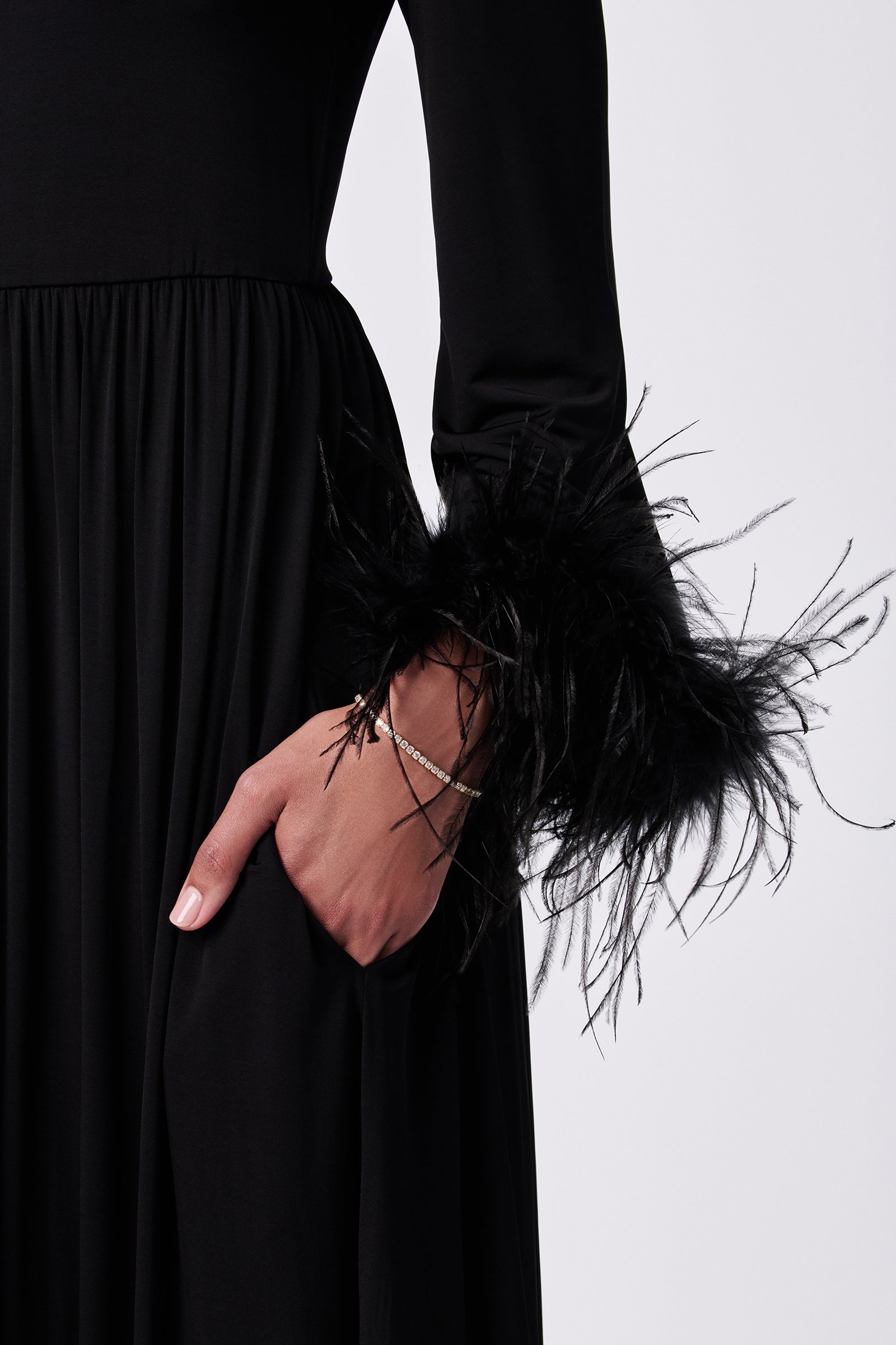 Wearing the Orla Feather-Cuff Maxi Dress, featuring long sleeves and feathery black cuffs, a person with light pink nails and a delicate bracelet poses with one hand in their pocket. The dress also has a gathered waist.