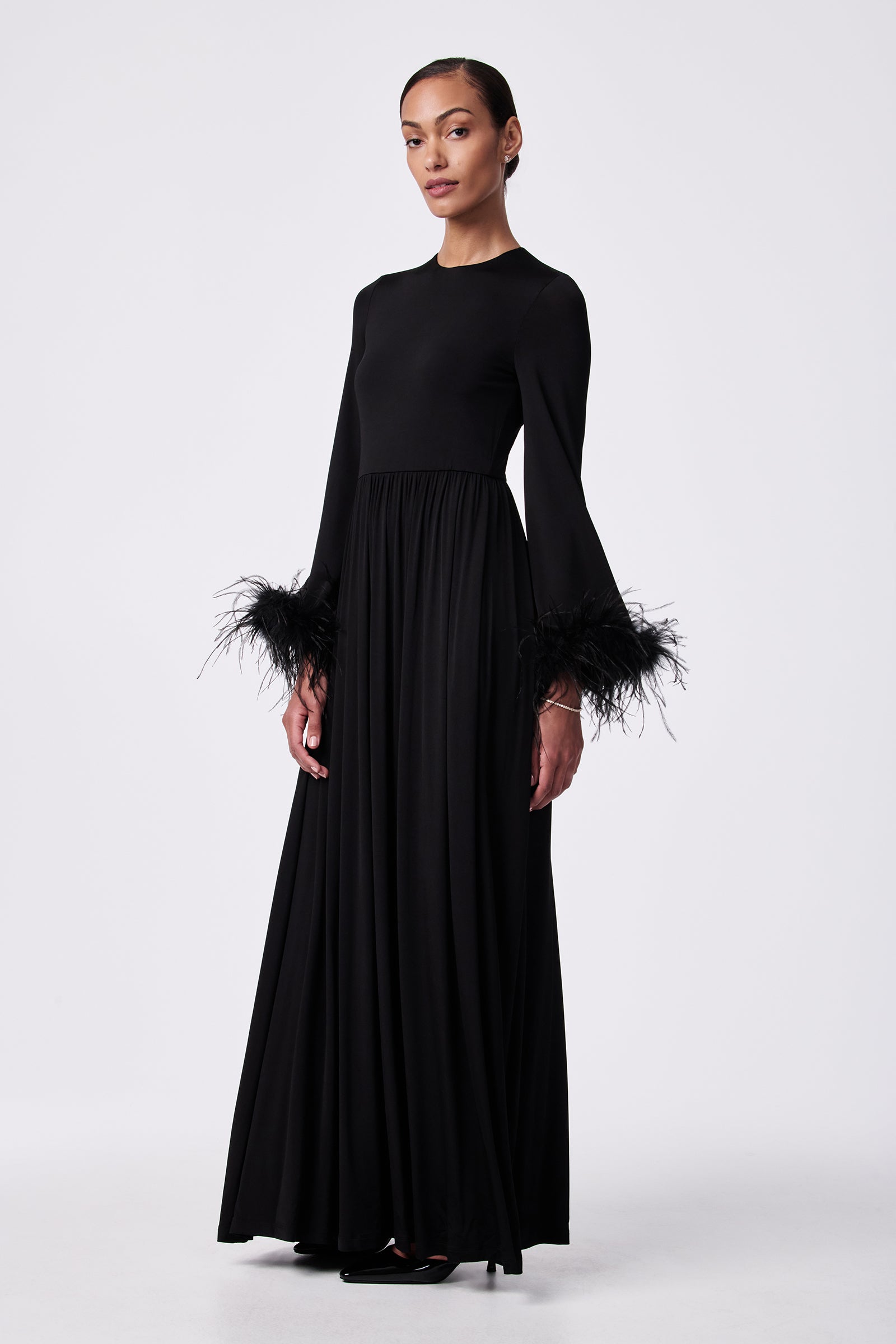 A woman wears the Orla Feather-Cuff Maxi Dress, a long black gown with long sleeves and black feather details at the cuffs, standing against a plain white background.